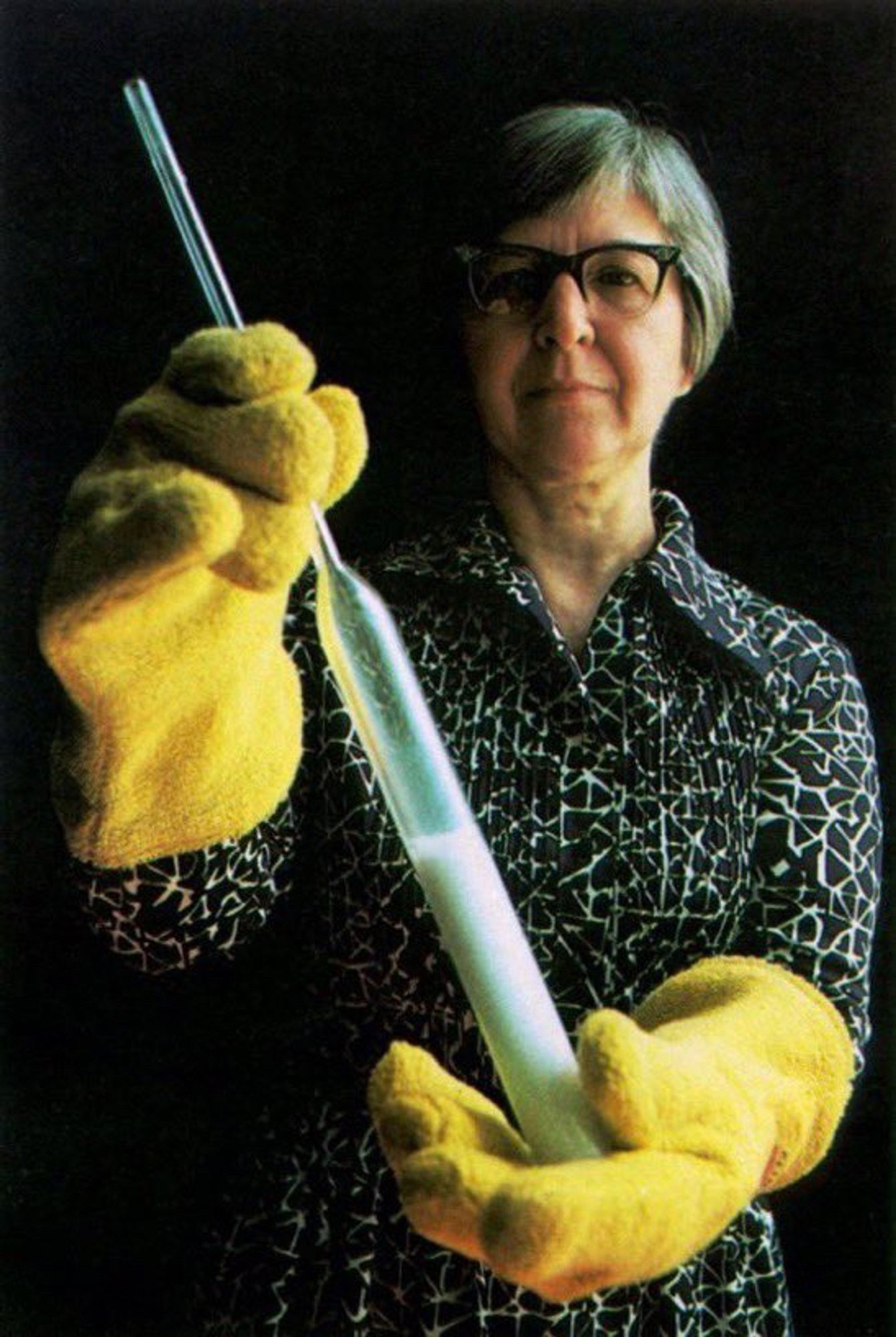 Color photo of Stephanie Kwolek. She wears a blouse with a batik patter. Her hair is still in a short bob, but is graying. Kwolek wears large, yellow rubber gloves and is holding a large pipette full of milky white fluid up to the camera.