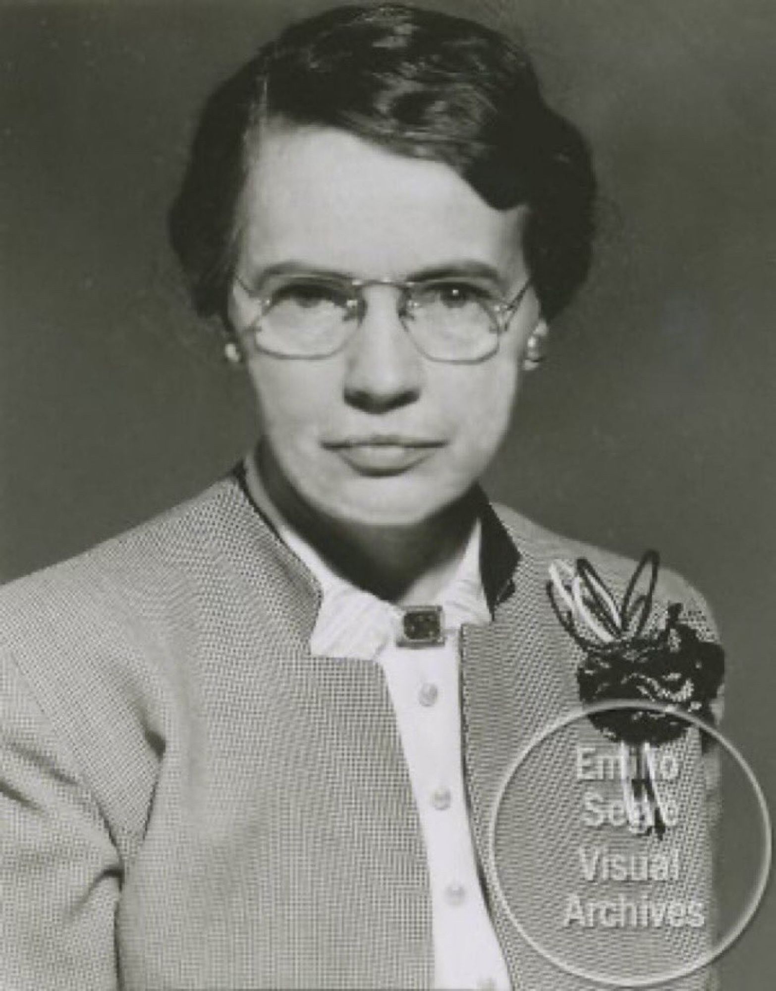 Black and white photo of Charlotte Moore Sitterly. She has short hair, glasses, and is waiting a houndstooth coat and white blouse fastened with a broach. She is looking directly at the camera.