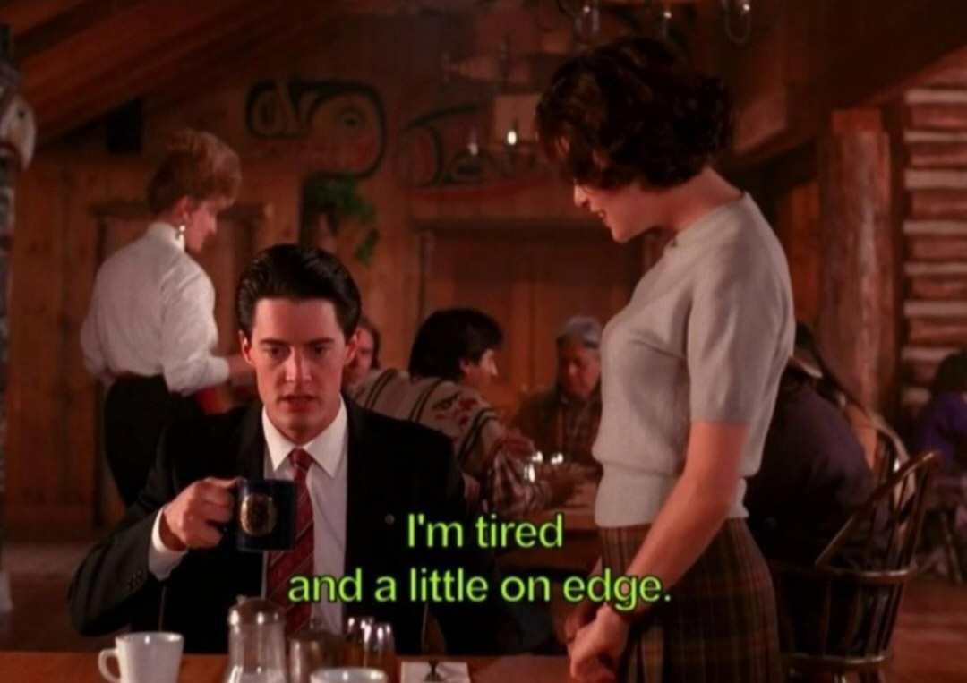 A still shot from Twin Peaks, in the dining room of the Great Northern Hotel. FBI Agent Dale Cooper is sitting at a table lifting a cup of coffee to take a sip. Audrey Horne is standing to the right and other diners are visible in the background. Coop is saying: 
"I'm tired and a little on edge."