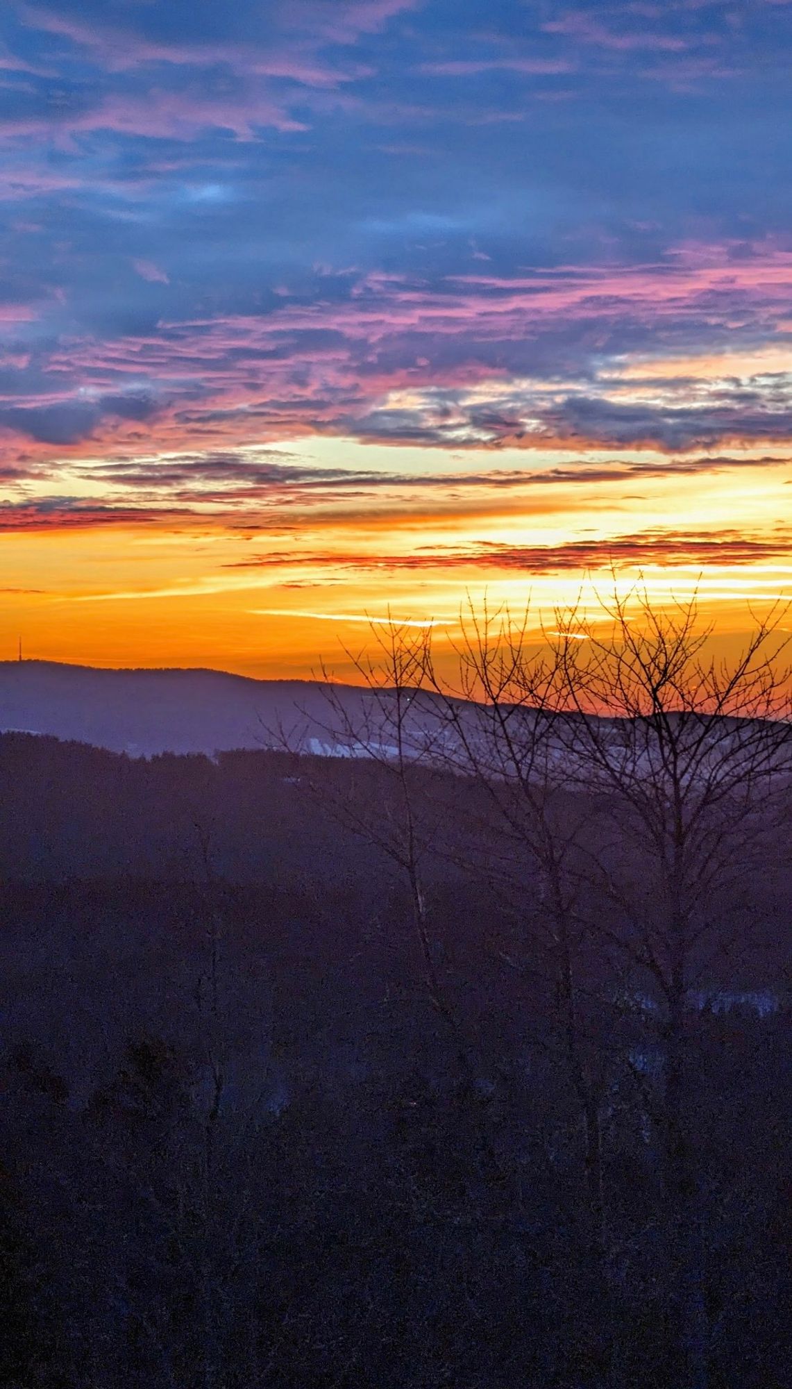 Das Bild zeigt einen wunderschönen Sonnenuntergang über einer hügeligen Landschaft. Der Himmel ist in kräftigen Farben von Orange, Gelb, Lila und Blau getaucht. Im Vordergrund sind Silhouetten von kahlen Bäumen zu sehen, die die dunklen Hügel betonen. Die Szenerie wirkt ruhig und friedlich, während die Farben des Himmels einen atemberaubenden Kontrast zur Dunkelheit der Landschaft bieten.