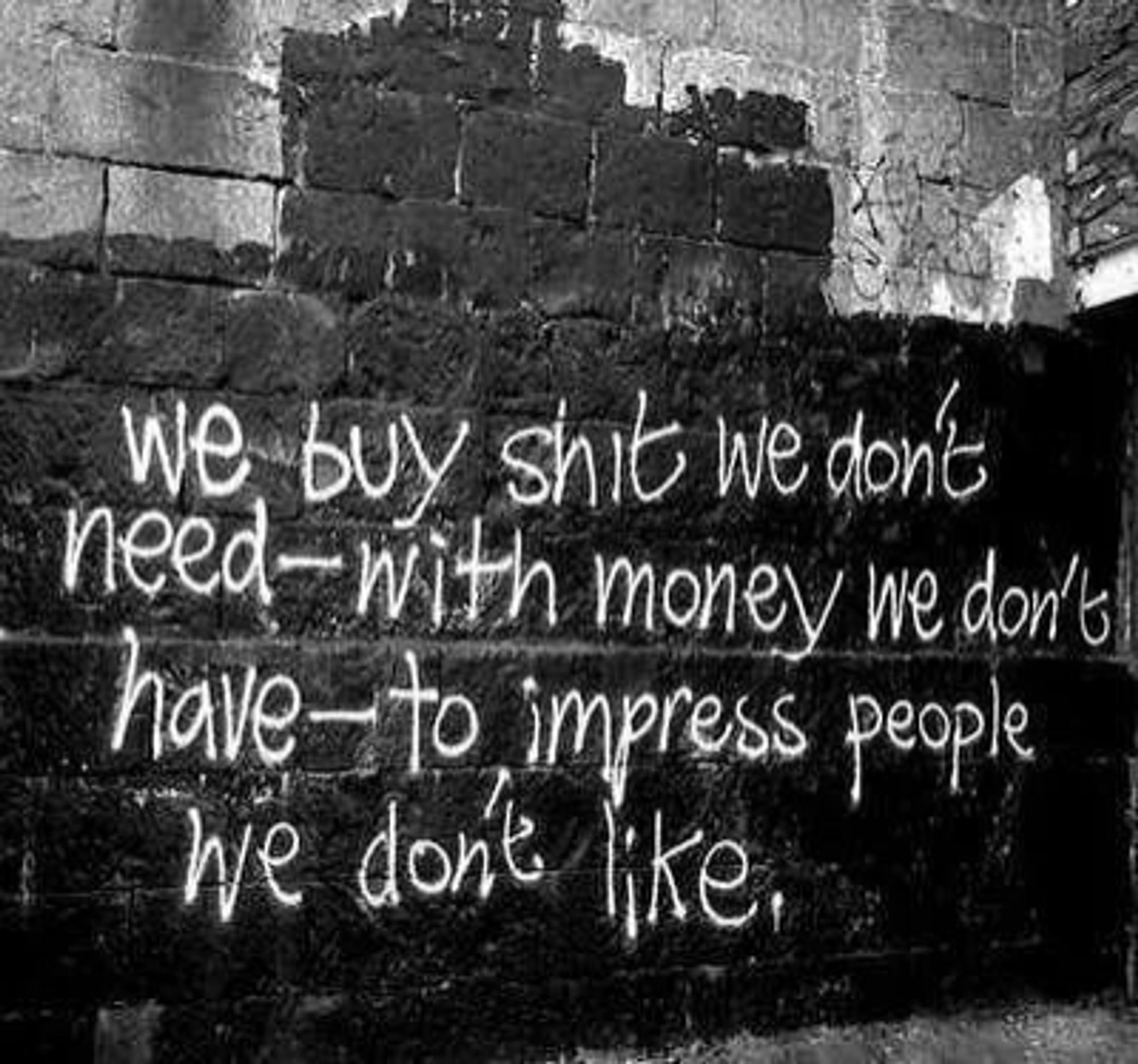 Auf einer Ziegelwand steht in weißer Schrift
We buy shit we don't need-with money we don't have-to impress people we don't like