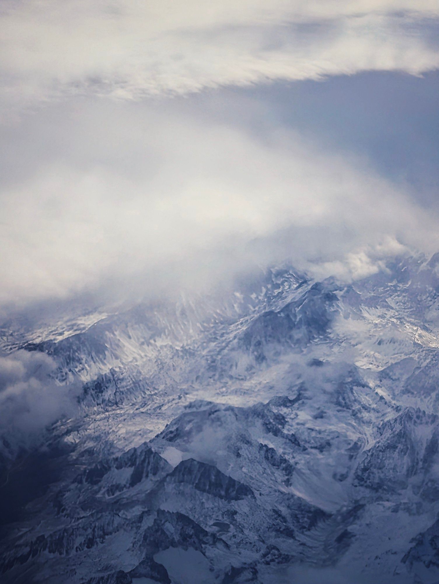 Die Alpen aus der Vogelperspektive fotografiert. Oben im Bild eine Wolkendecke, darunter einige Bergspitzen auf denen Schnee liegt.