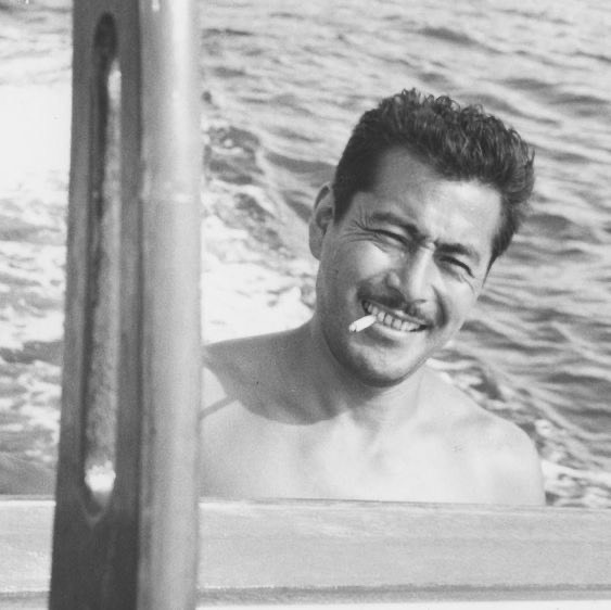 shirtless Toshiro Mifune piloting a boat , smiling with a cigarette between his teeth