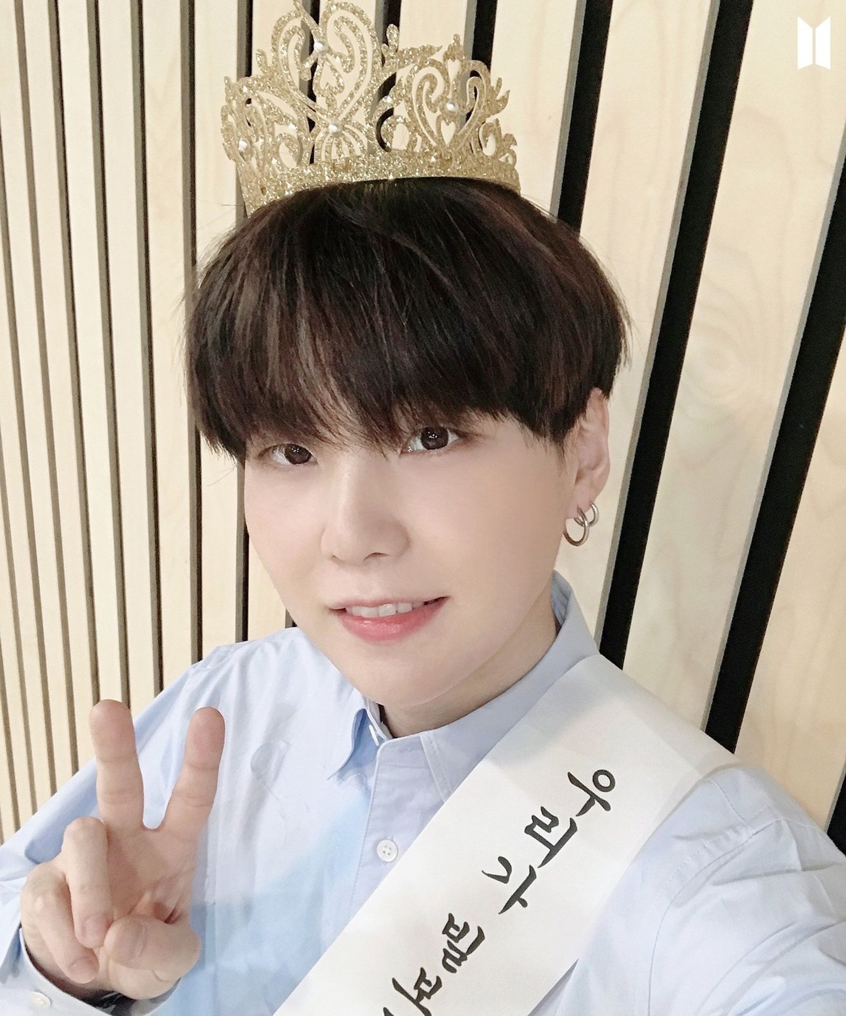 Yoongi wearing a crown and a sash with Korean letters, he has a blue long sleeve shirt and black hair. He looks adorable.