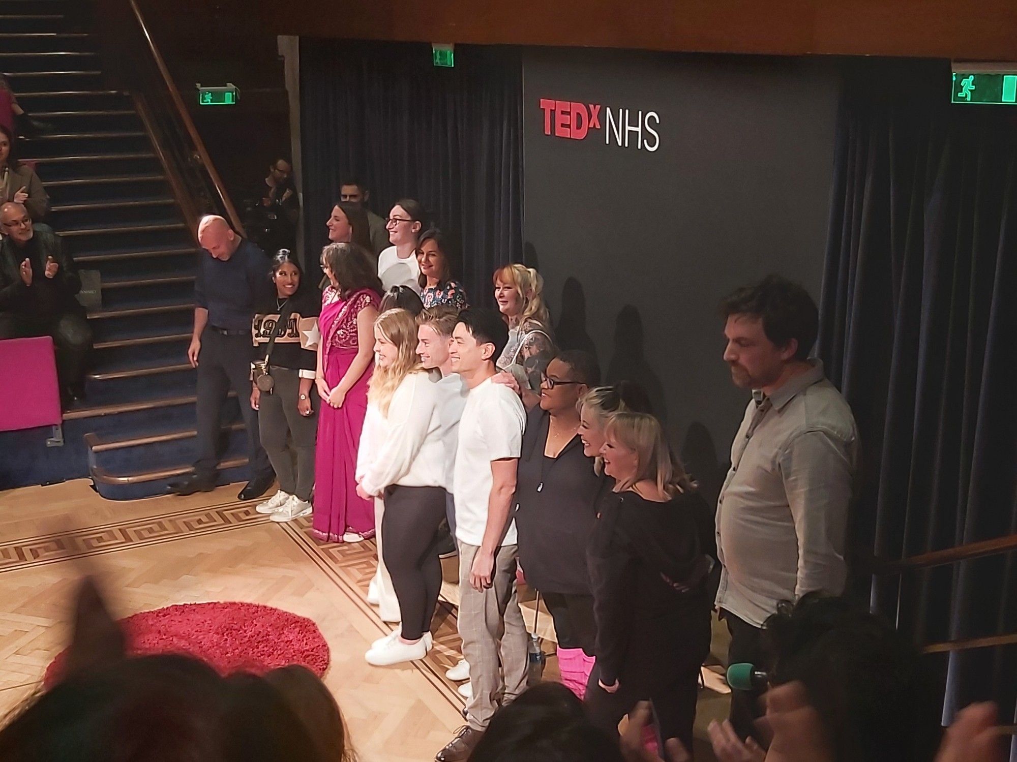 All the speakers of TEDxNHS as they stand in front of the black backdrop with the red and white logo.