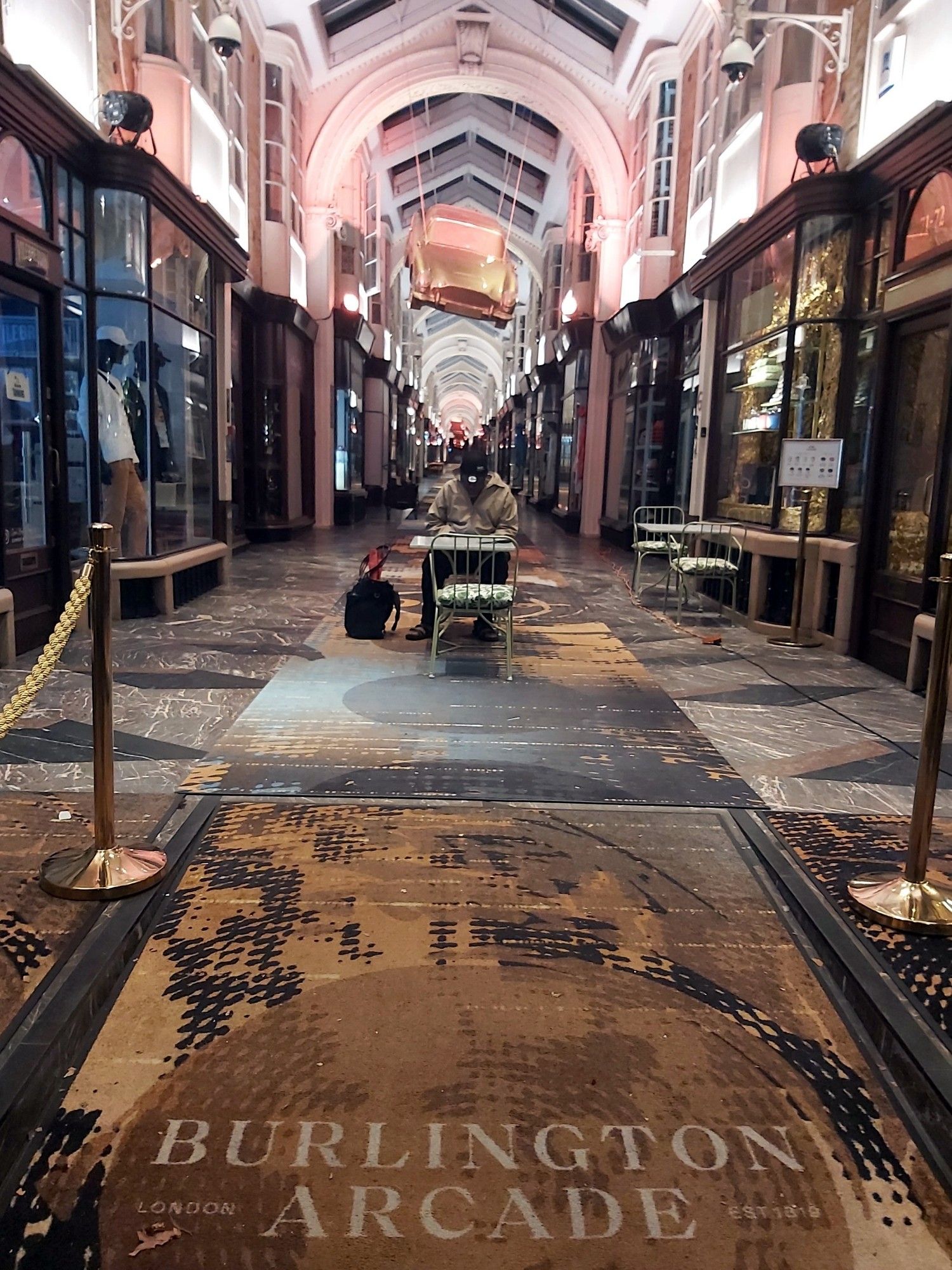 A posh shopping arcade with a figure in the foreground seated at a table. The nearer foreground has the words "Burlington Arcade" in serif capitals.