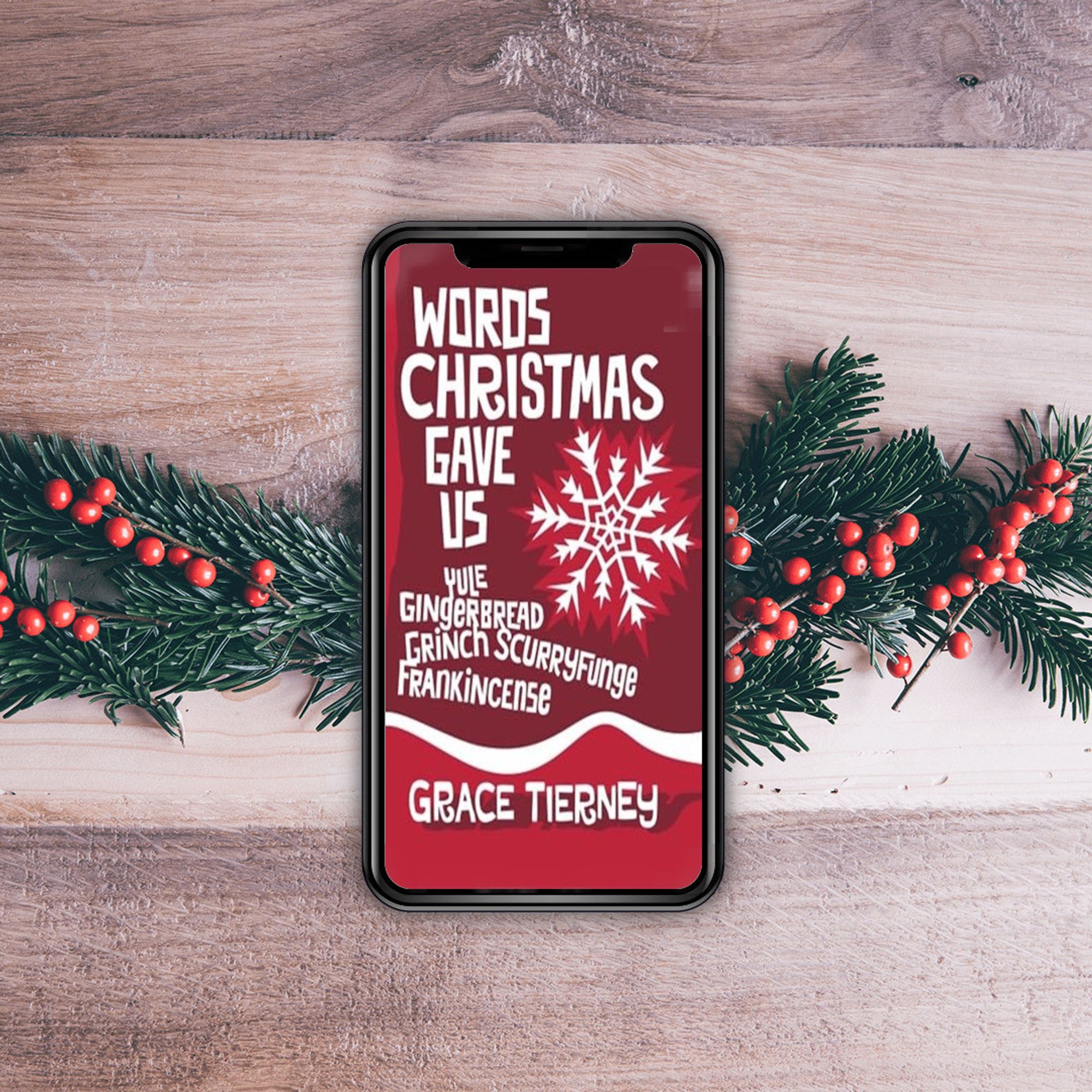 Cover of "Words Christmas Gave Us" on a mobile phone on top of a Christmas foliage garland on a wooden background.