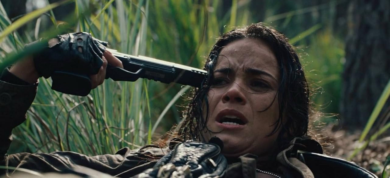 A still from Predators (2010) in which Braga's character Isabelle points a pistol at her own head while lying on the jungle floor.