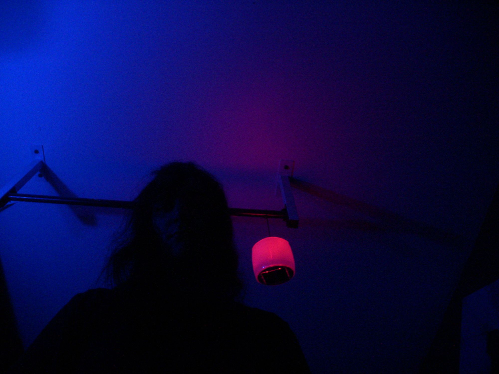 A photo of a young woman looking at the camera from overhead, her features completely silhouetted by darkness other than vaguely definable facial features. The room is dimly lit by a deep blue ambient light and the glow of a red lantern hanging to her right.