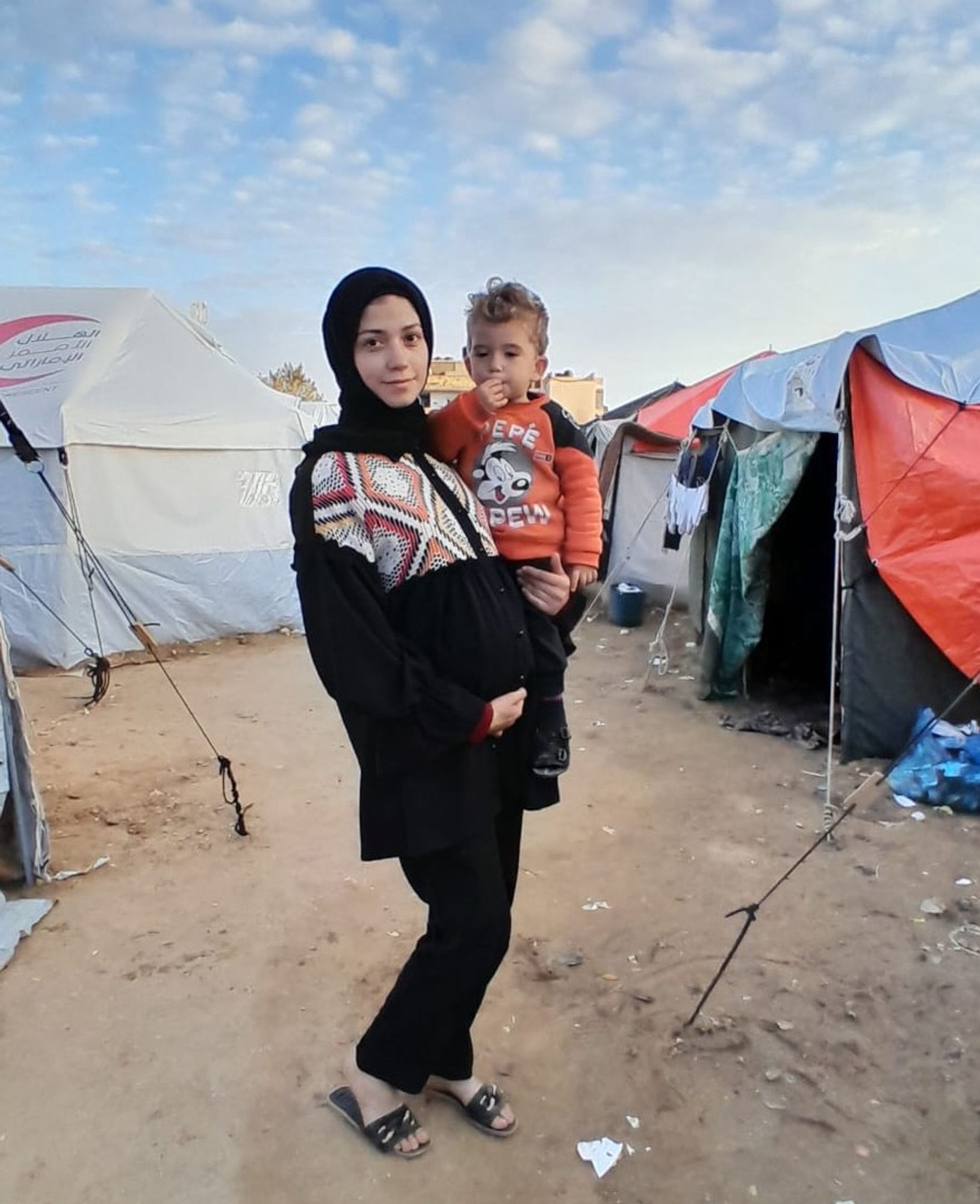 A pregnant Lama Zaqout stands with her son in her arms surrounded by a blue cloudy sky and tents in Rafah, Gaza.