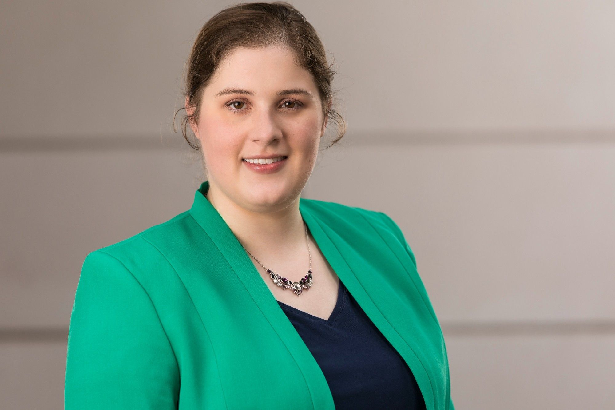 Sarah, a white woman with brown hair pulled back, smiling at the camera. She is wearing a green blazer and navy blouse.