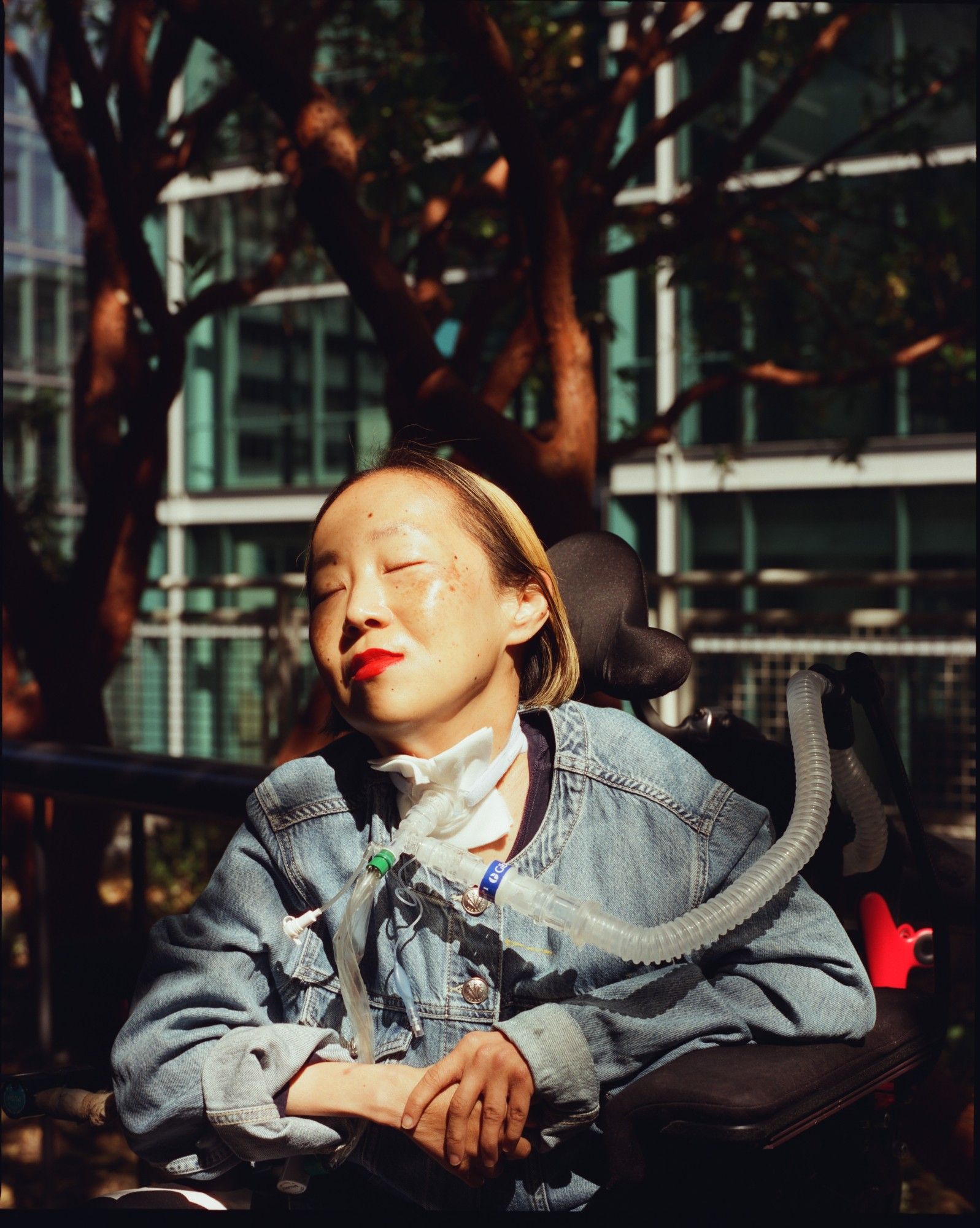 An Asian American disabled woman is sitting in a wheelchair. She has blonde highlights in her hair and is wearing a denim jacket. She has a bright red lip color on and a tracheostomy at her neck connected to a ventilator tube. Her eyes are closed her face has a dreamy expression. The sun is shining over her while the background of trees and a skyscraper is shrouded in darkness. Photo credit: María del Río