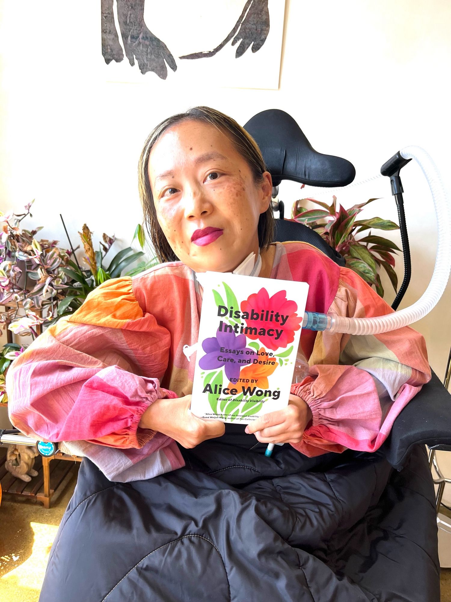 Asian American woman in a wheelchair with a tracheotomy at her neck connected to a ventilator. She is wearing a plaid shirt with pastel colors and a magenta lip color (Chanel Milky Blueberry). Behind her are a bunch of plants and a bottom half of a poster of a frog.