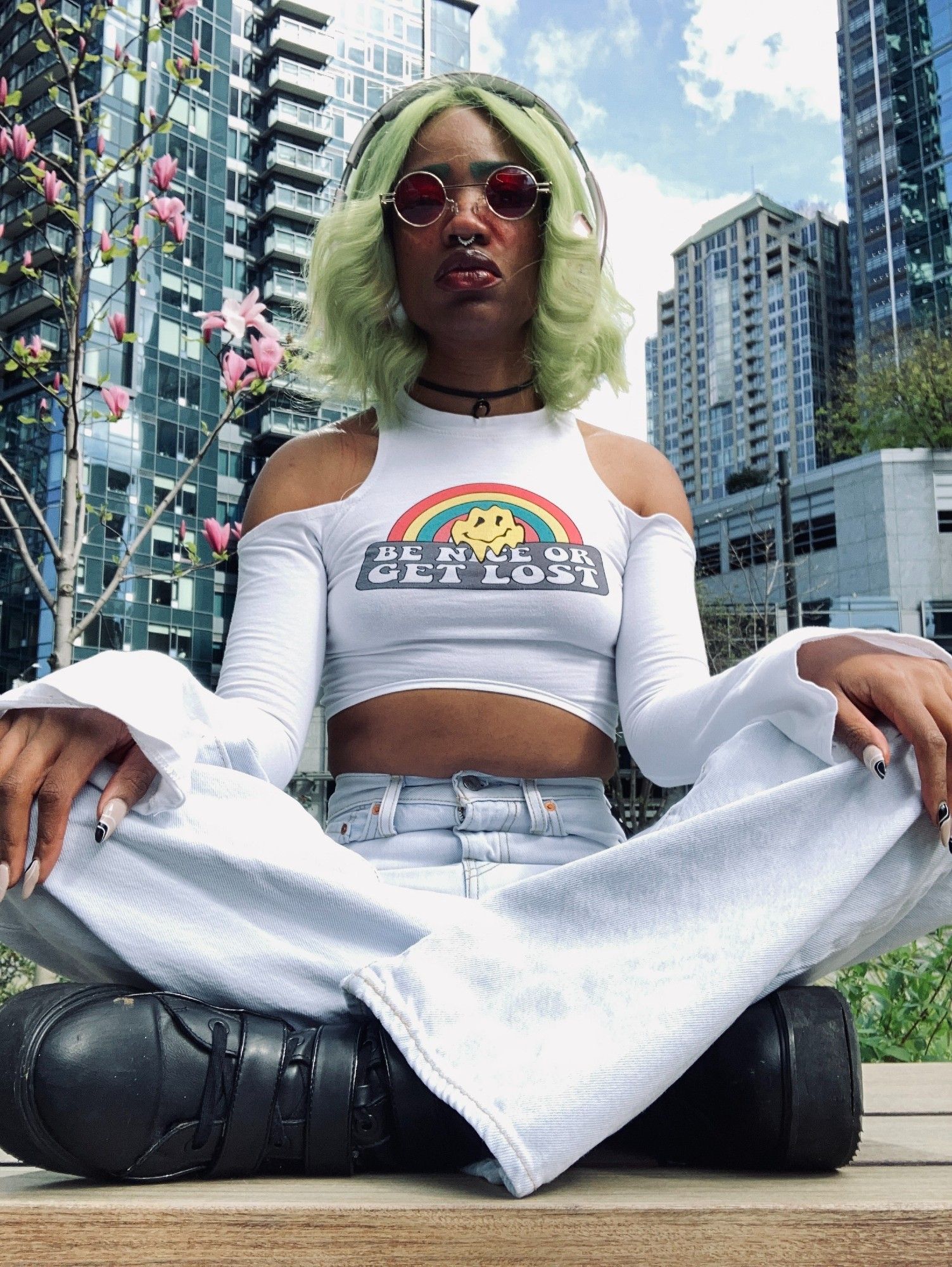 Image of Faati, a nonbinary light brown skinned Black Native, sitting cross legged in front of a cherry blossom tree and multiple high rise buildings. They have on acid washed light blue bell-bottom jeans, a white crop topped long sleeved shirt  and black platform boots. They are sitting cross-legged with their hands on their knees. Their fingernails are a long almond acrylic style with nude coloring and black and white designs on top. They have green straight 70s styled hair with the ends bumped. They are wearing circle 70s style red lens sunglasses.