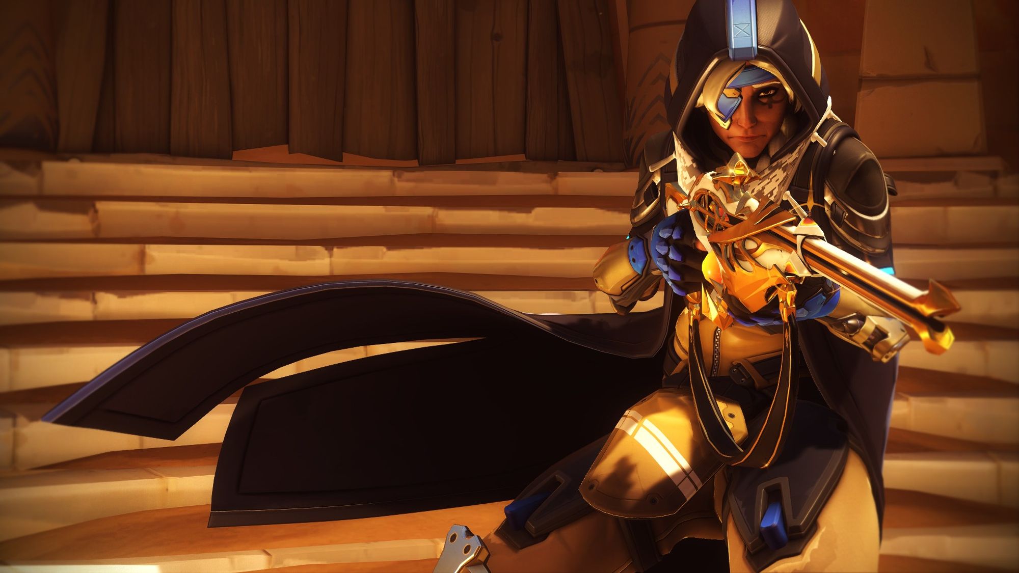 Overwatch 2 screenshot of Ana in her basic Overwatch 2 outfit. It is a tactical suit with a long blue duster with hood, a scarf that looks similar to a keffiyeh, an eye patch and a hijab under her hood. Her suit underneath is beige. She has a very ornate white and gold-detailed sniper rifle with crystal details. The shot is with her looking straight into the camera, holding her gun aloft. Her duster is floating in the wind as if she just moved.