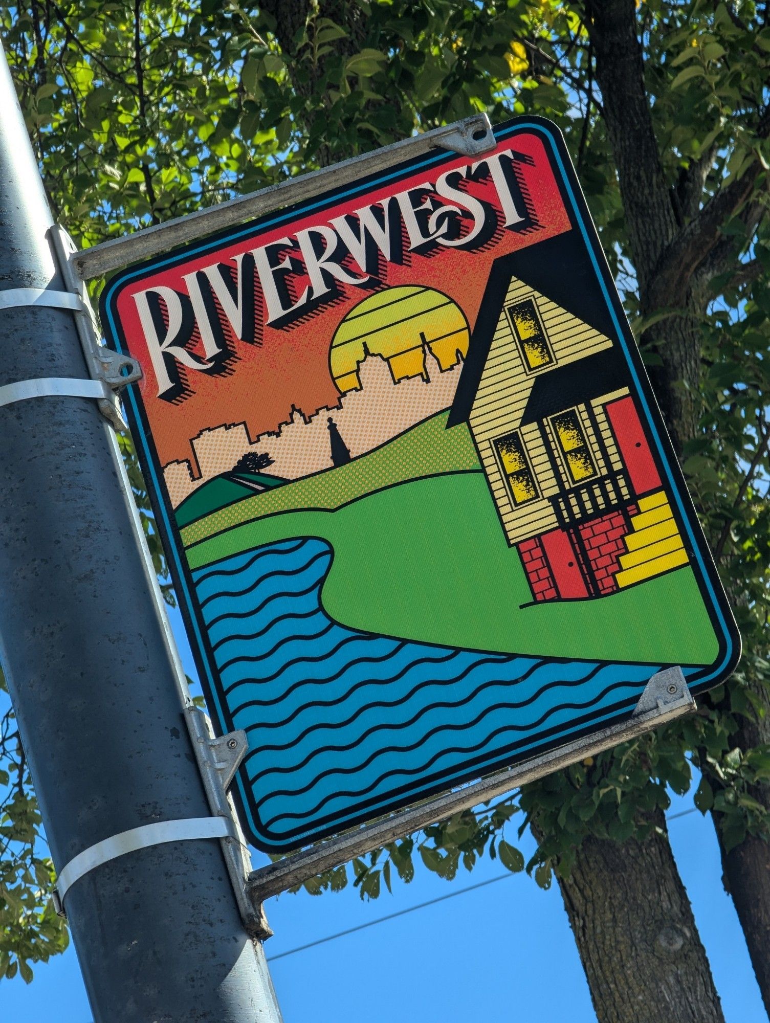 Street sign indicating the Riverwest neighborhood; it is in a vintage 70s style with flat colorful designs of a river and house next to it with the setting sun behind a skyline of downtown Milwaukee