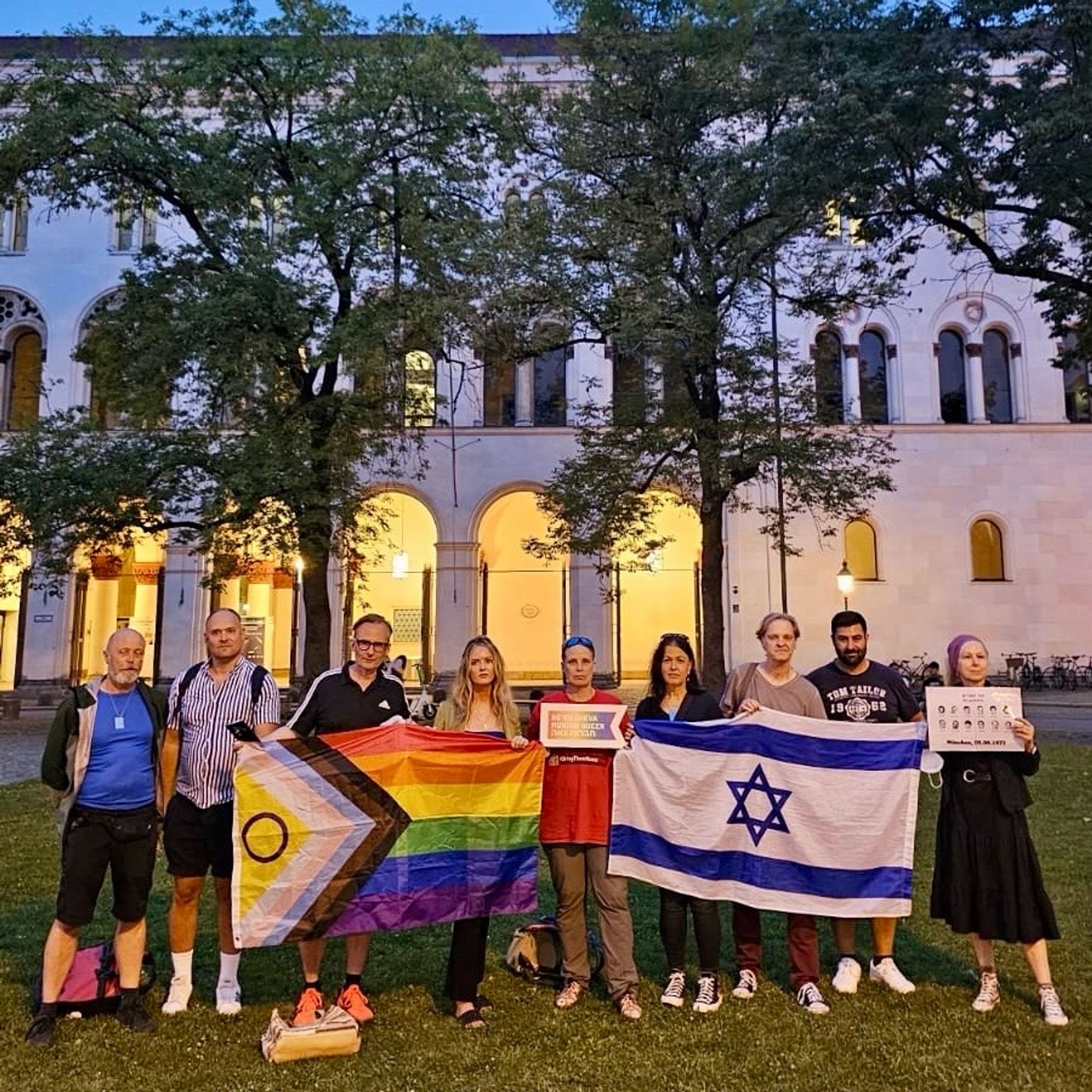 Das Bild zeigt eine Gruppe Menschen vor der abendlich beleuchteten Münchner Universität am Geschwister-Scholl-Platz. Sie halten die Pride Flag, eine Israelflagge und ein kleines Schild des städtpartnerschaftlichen Projekts Be‘er Sheva Munich Queer.