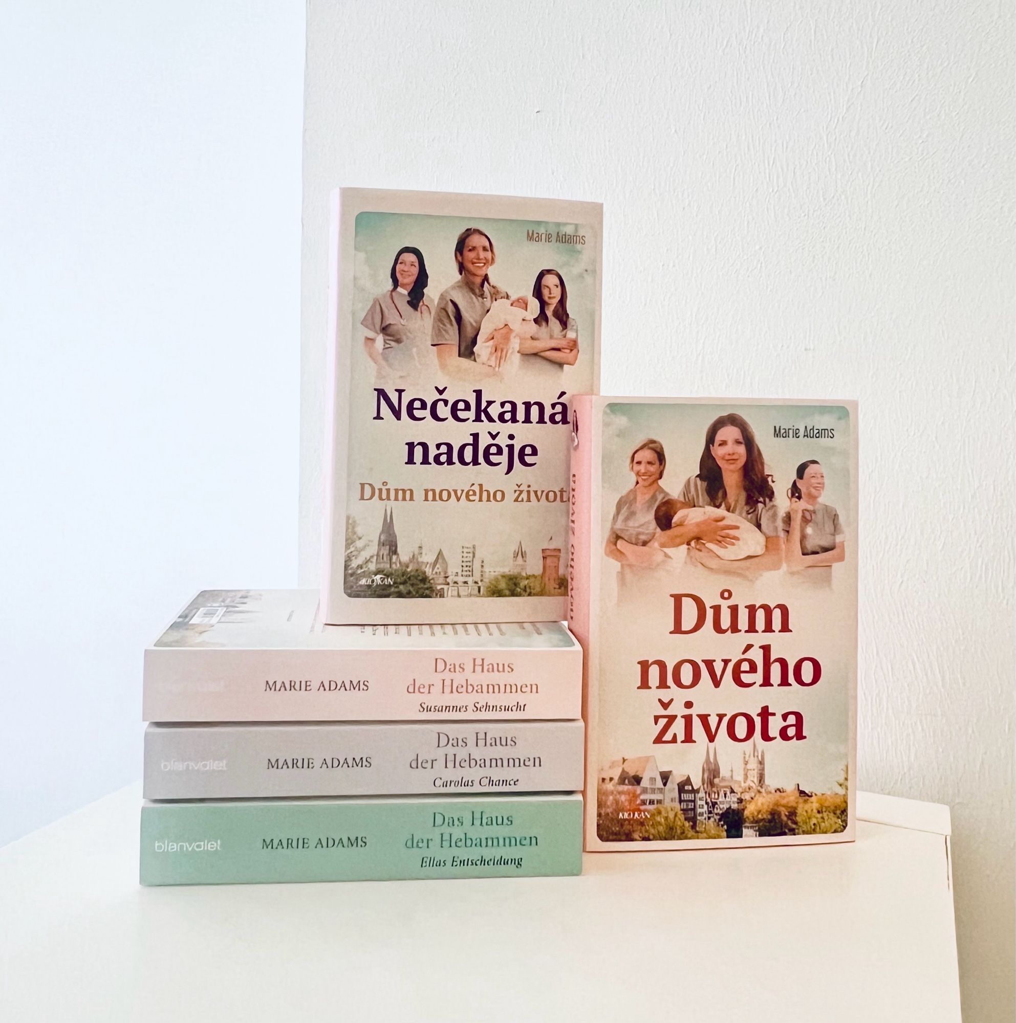 Das Bild zeigt einen Stapel Bücher vor einem weißen Hintergrund, drei in deutscher und zwei in tschechischer Sprache. Die beiden tschechischen Bücher stehen aufrecht und sind auf der Vorderseite zu sehen, links der Titel »Nečekaná naděje: Dům nového života«, rechts »Dům nového života«, beide von Marie Adams. Die Cover zeigen drei Hebammen, die ein Baby in den Armen halten, mit einer Stadtansicht im Hintergrund. Unter den tschechischen Ausgaben liegen drei deutschen Originalausgaben der Reihe »Das Haus der Hebammen«: Susannes Sehnsucht, Carolas Chance, Ellas Entscheidung. Die deutschen Bücher haben ein ähnliches Coverdesign.