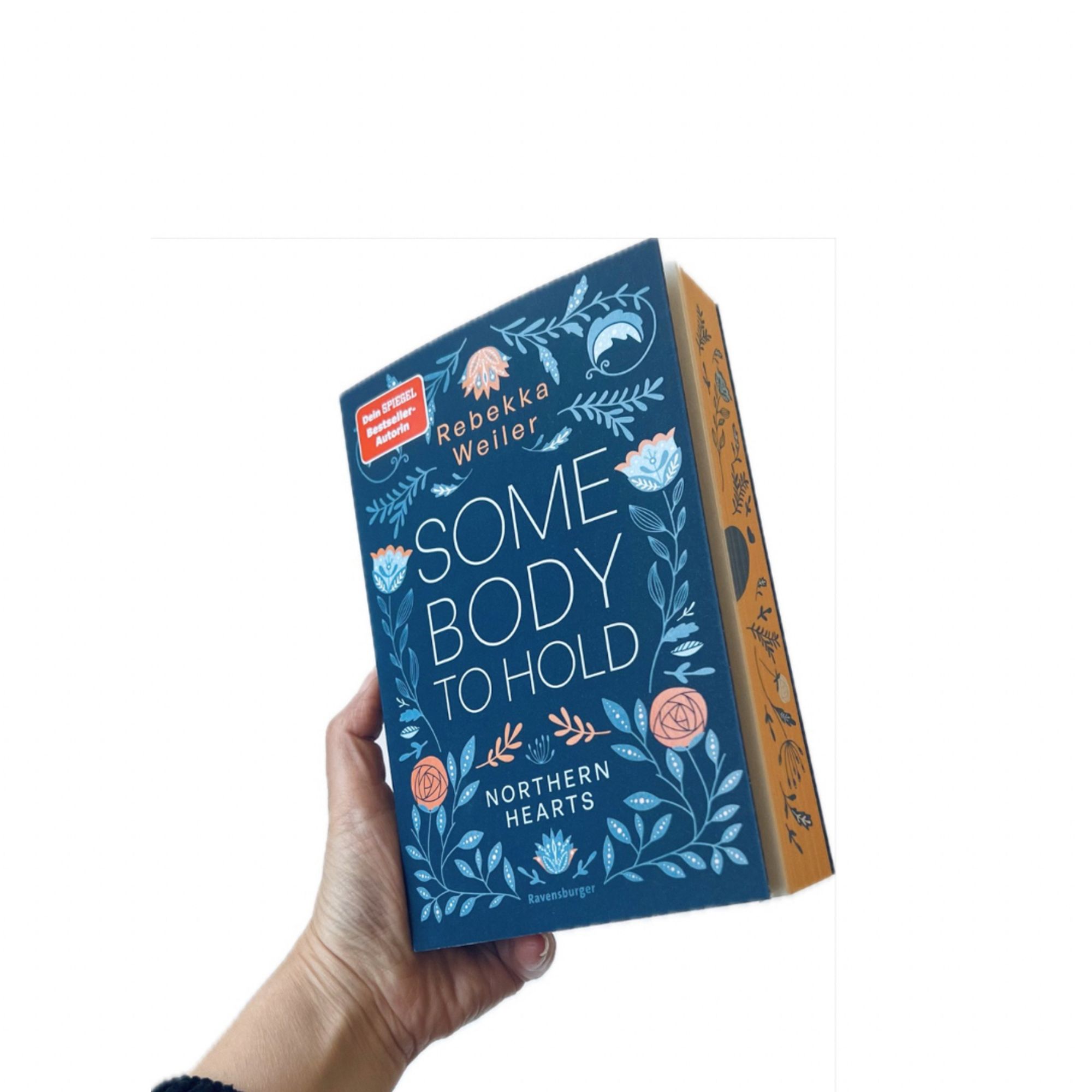 Eine Hand hält ein Buch mit dem Titel »Somebody to Hold« von Rebekka Weiler. Das Cover ist blau mit floralen Verzierungen. Auf der Seite des Buches sind ähnliche Blumenmuster in Orange zu sehen.