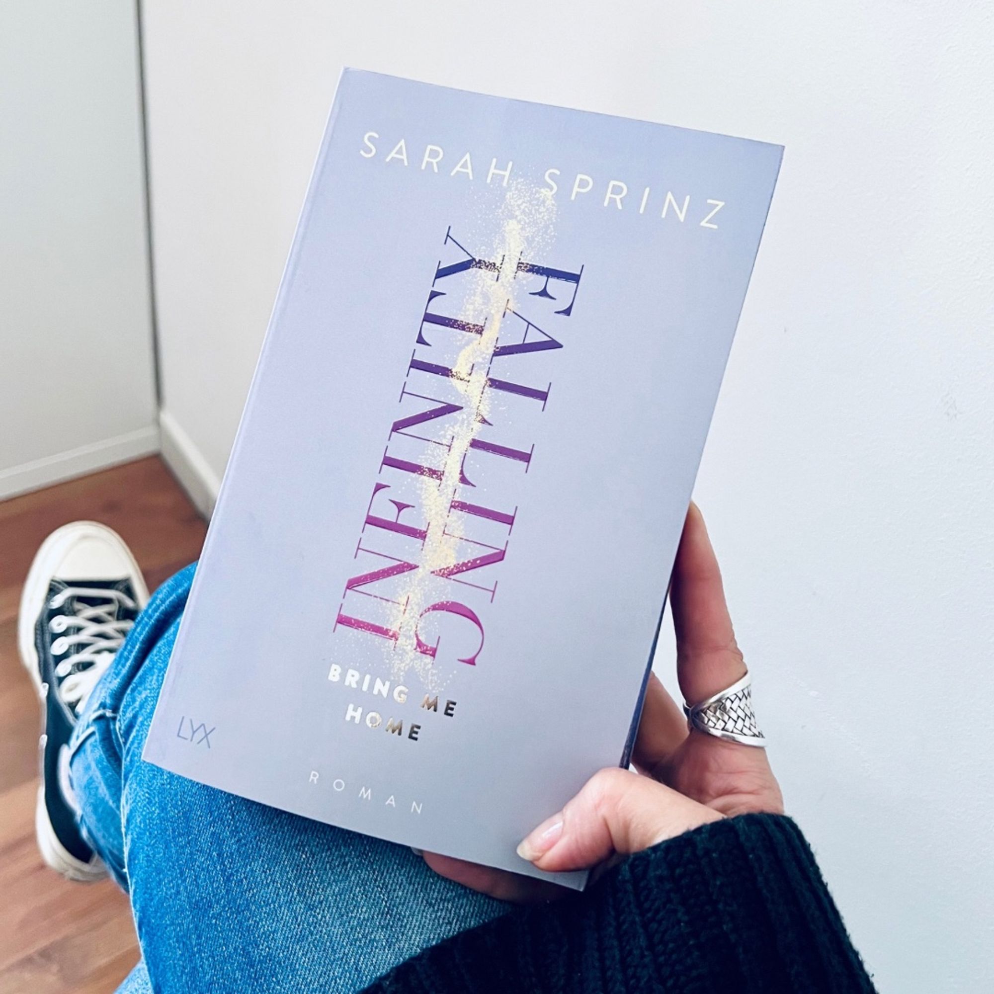 Auf dem Bild hält eine Person in Jeans, schwarzen Chucks und schwarzem Wollpulli das Buch »Infinity Falling: Bring Me Home« von Sarah Sprinz. Das Buch hat ein pastelllilafarbenes, grafisch gehaltenes Cover, erschienen ist es bei LYX.