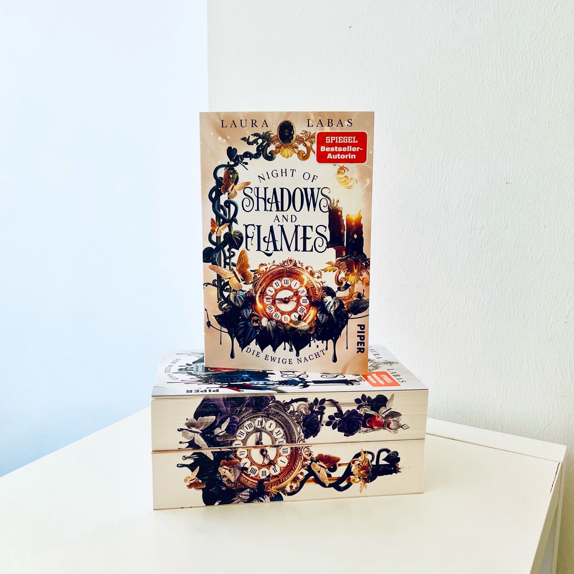 Das Bild zeigt das Buch »Night of Shadows and Flames: Die ewige Nacht« von Laura Labas. Es steht auf zwei weiteren liegenden Büchern (Band 1 und 2 dieser Dilogie). Die Cover sind kunstvoll gestaltet, beide aufeinanderliegenden Farbschnitte ergeben zusammen eine Taschenuhr.