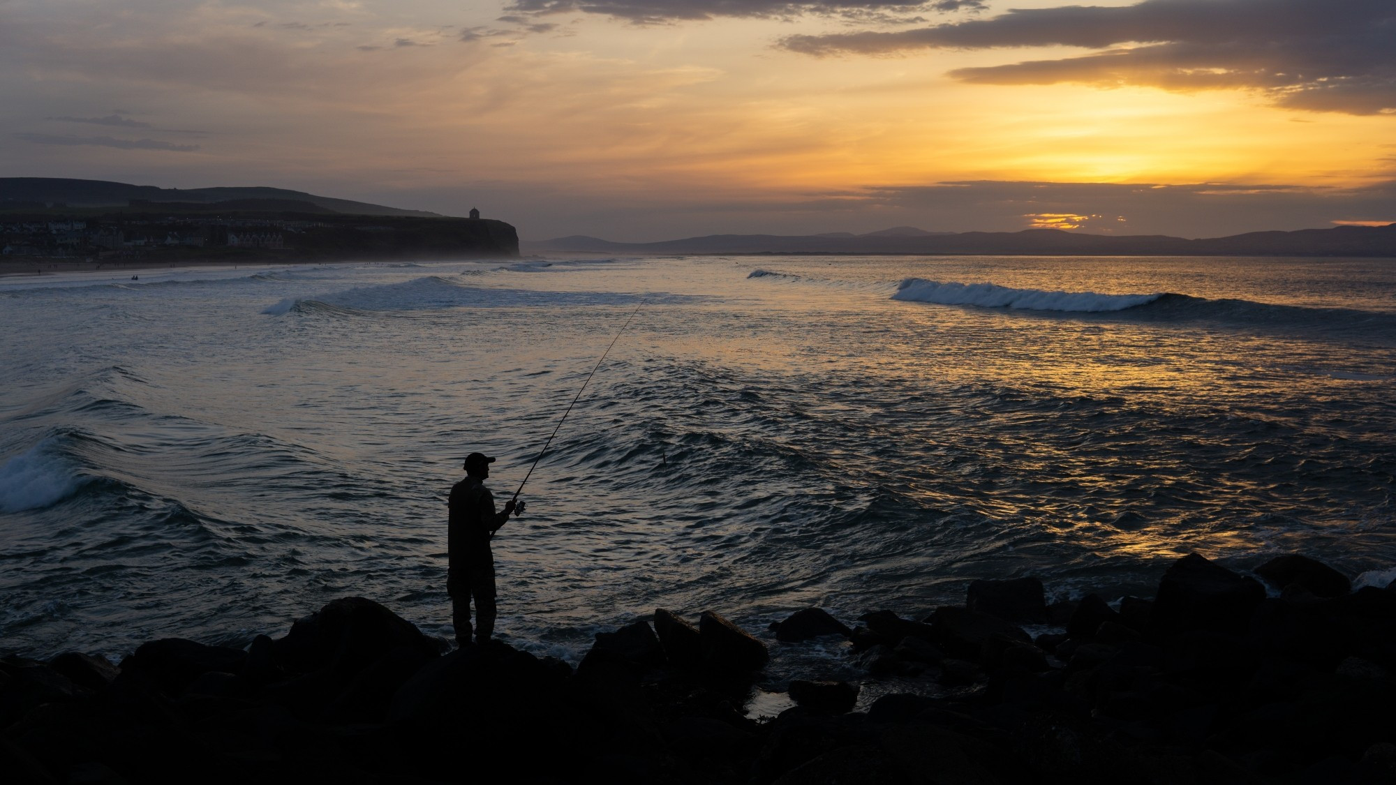 A landscape photograph taken at sunset. The sun has just set to top right of shot, and the light of the setting sun is reflecting on the waves of the ocean between the foreground and the horizon. A headland juts out into the sea on left on shot. A fisherman casts his rod from a breakwater in the foreground.