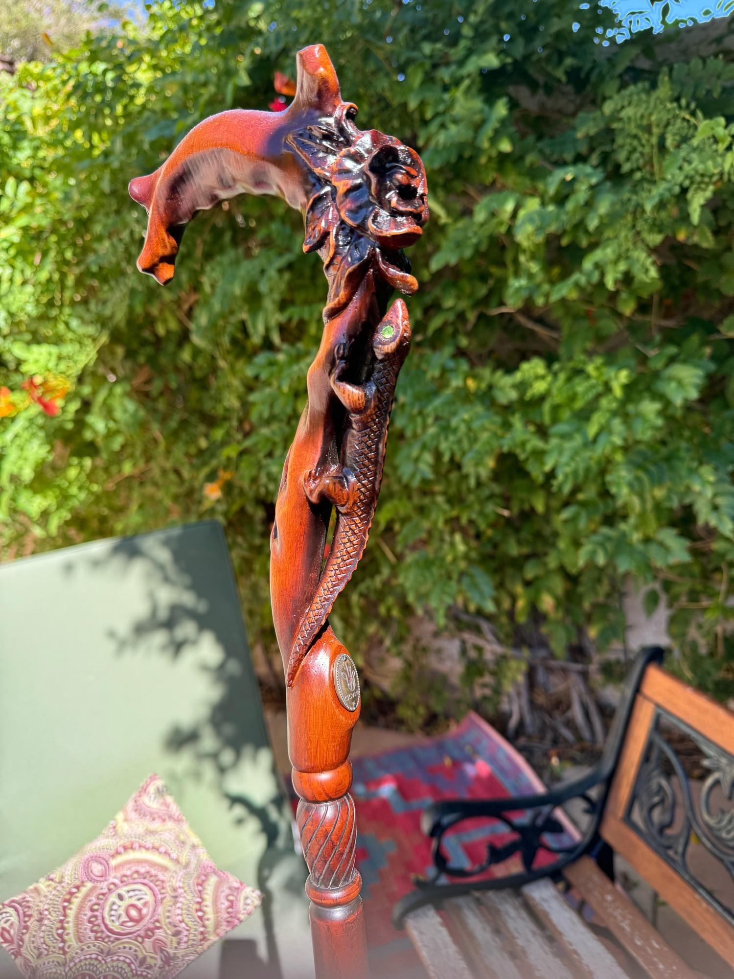 An ornately carved wooden cane that features a lizard, and a rose in three dimensional relief on the front of it. Below the lizard is a brass medallion set into the cane. The lizard has green jewels for eyes. In the background is a green chaise lounge, a wood and iron bench And a trumpet vine with orange flowers. There is a brightly colored magenta and blue Oriental style rug on the concrete.