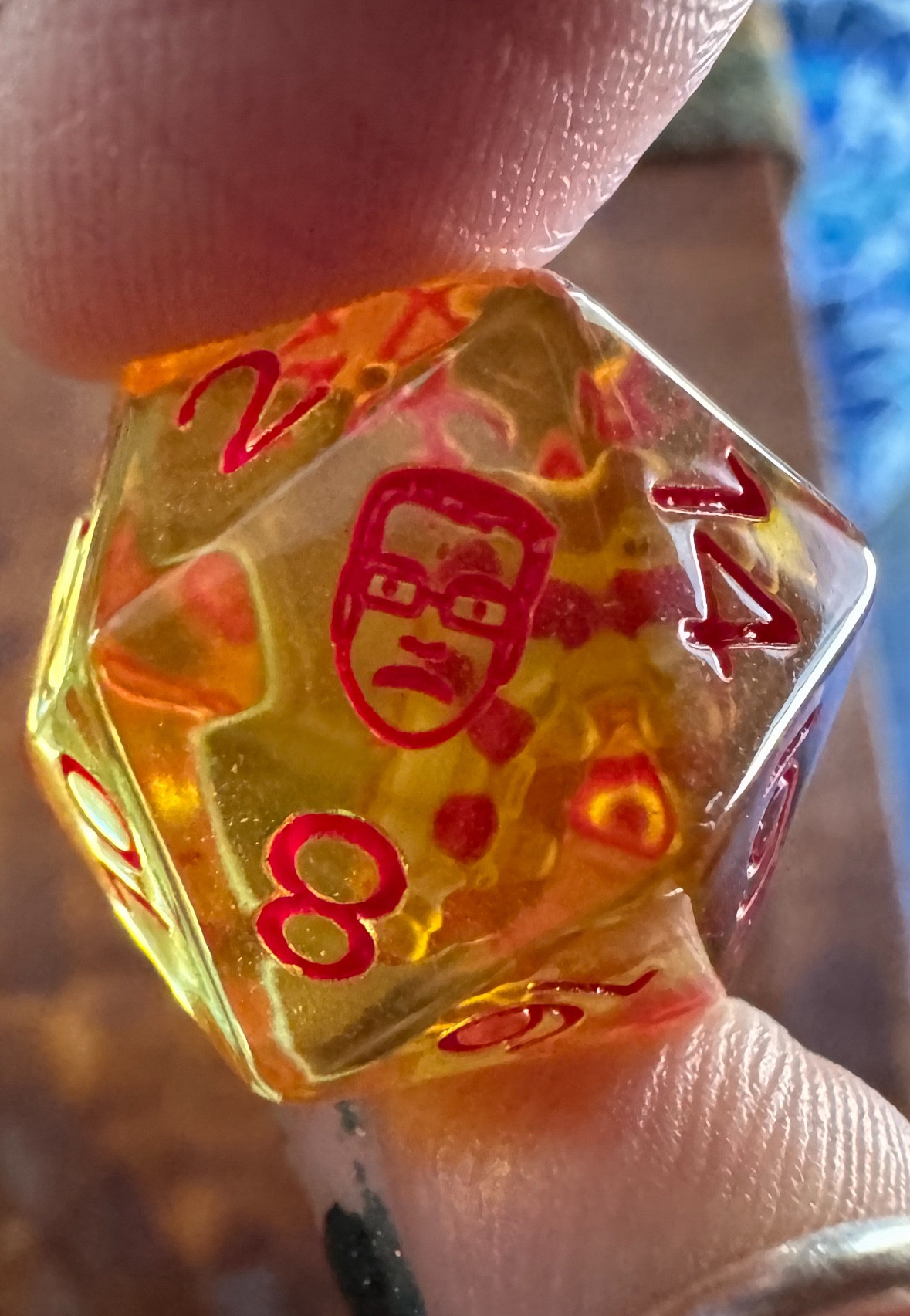 A close-up of the Pizza John d20 die, a transparent yellow dye with red numbering that features a little slice of pepperoni pizza inside. Pizza John’s face represents the 20.