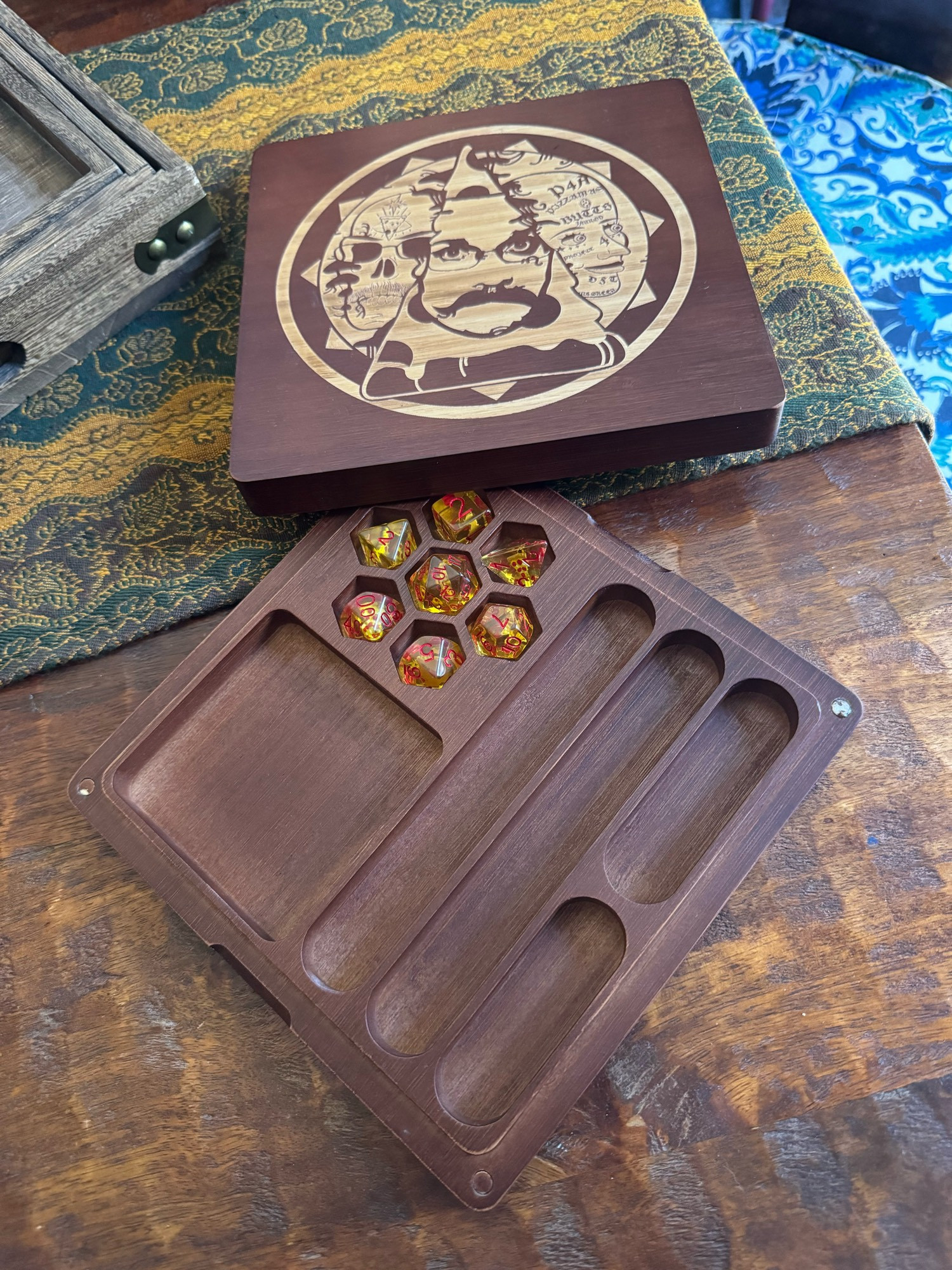 A dark brown wooden dice tray laying on a wooden table. The base of the tray contains multiple long slots, a square slot, an individual hexagonal dice slots, each each with a yellow polyhedral die in them. The lid features a circle around the main image. In the center is a slice of pizza standing up right like a pyramid with pizza John’s face in it. On the left is a skull wearing glasses that has a slice of pizza on its forehead topped by a star. On the right is a face covered in tattoos like”P4A” “pizzamas” “Butts Jarrod” “project” and “DST.” The image sits on a sun shape.