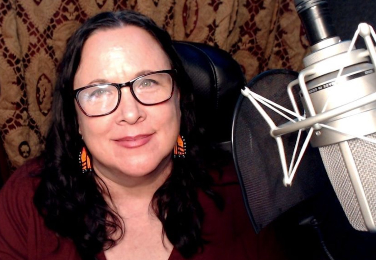 Selfie of me in my booth. I have wavy dark hair, hazel eyes and dark red glasses on. I’m wearing beaded earrings that each look like a monarch butterfly wing. My TLM-103 microphone is in the right foreground. Behind me are red and gold Indian-design curtains.