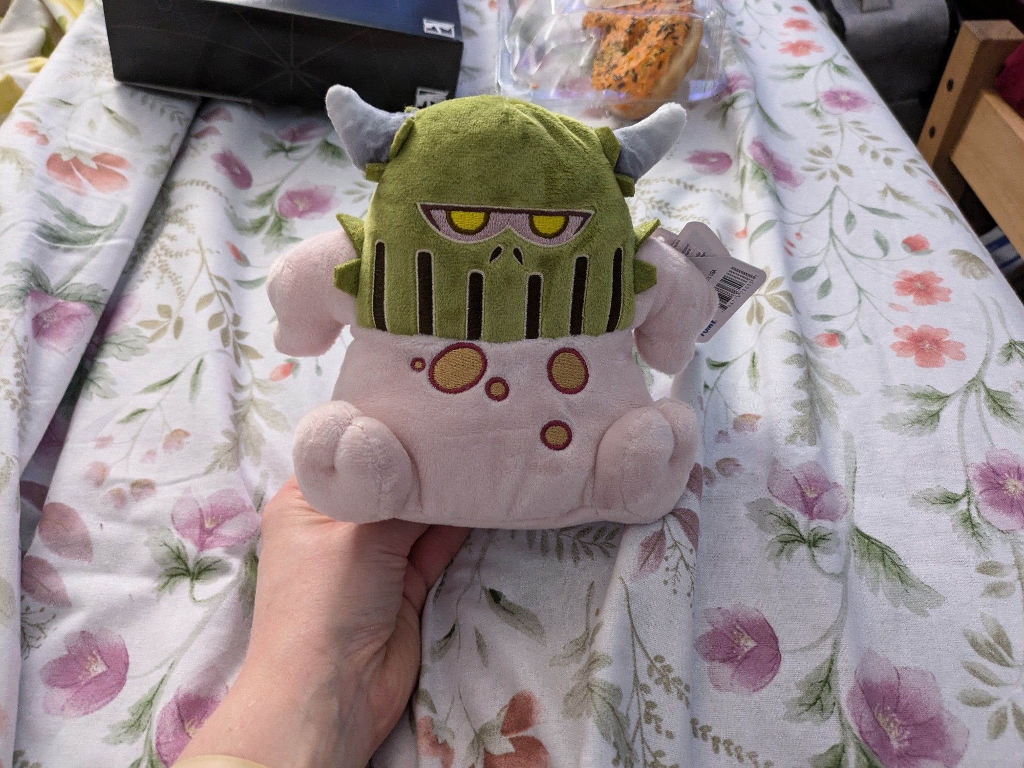 A plushie of a nurgling from Warhammer 40,000. This one has its hands on its hips and is wearing the helmet of a Plague Marine champion on its head.