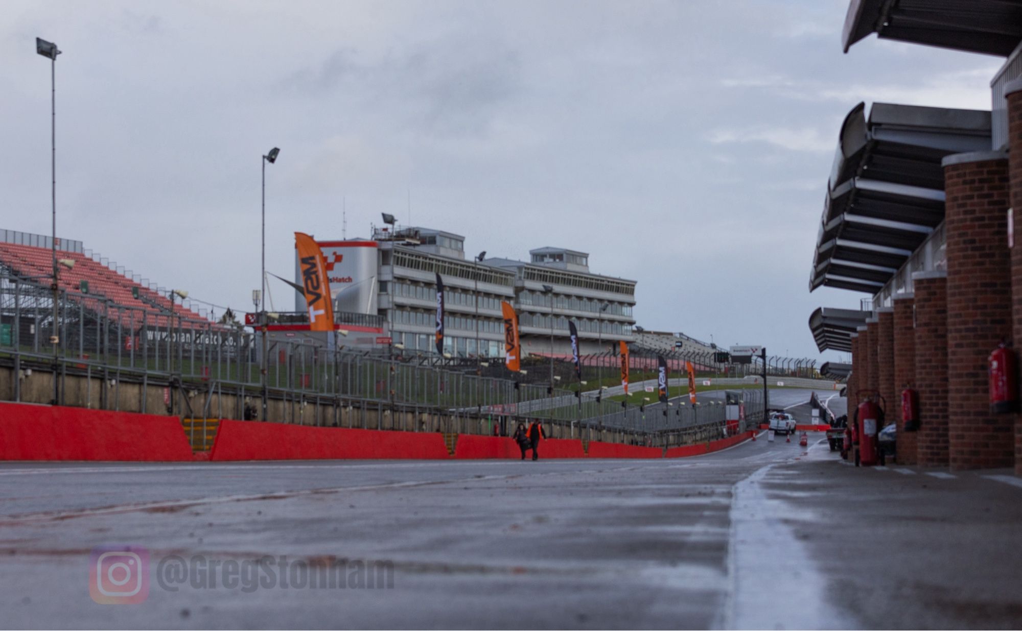 Rain falls on a near empty pit lane at Brands Hatch. Low angle wide shot towards the grandstands.