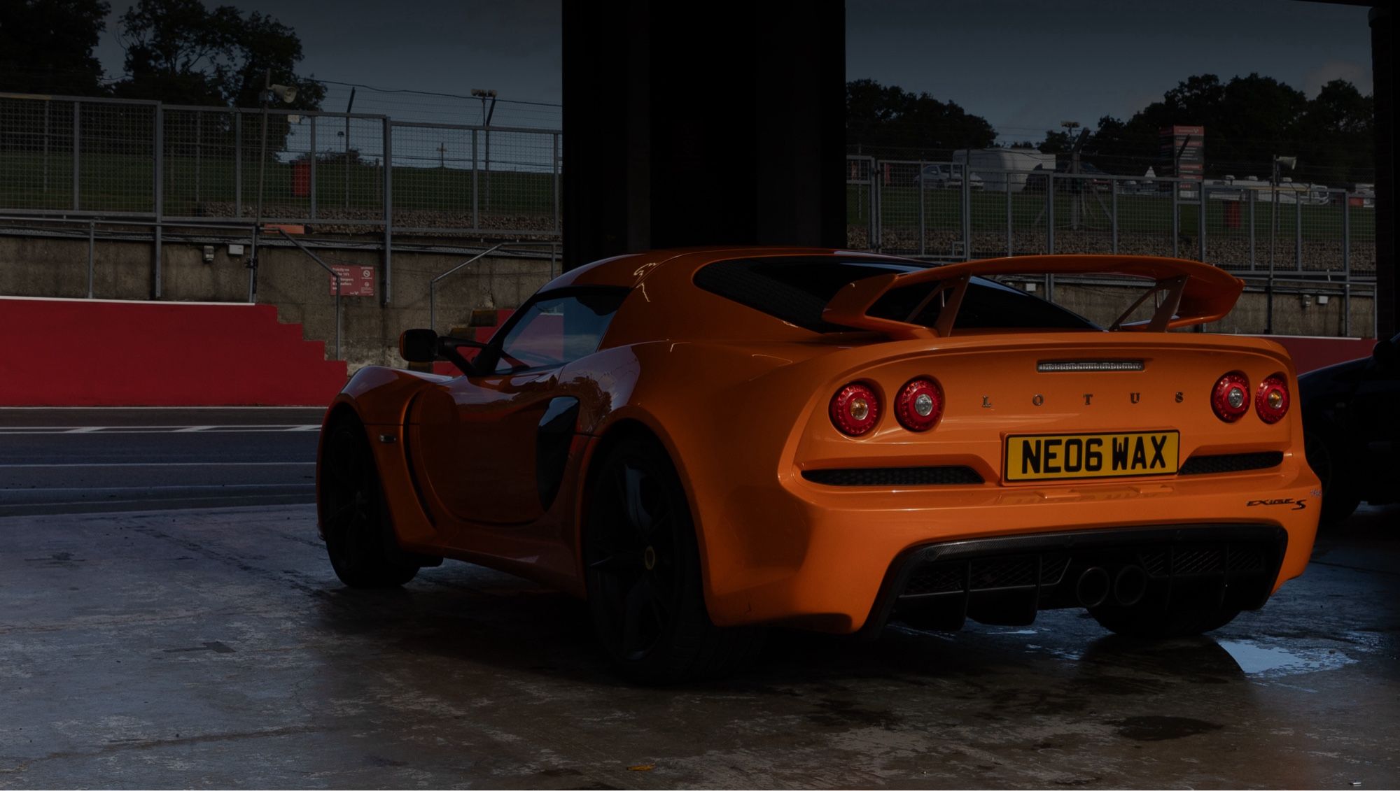 An orange Lotus Exige S parked in a pit garage at Brands Hatch. The rear of the car is illuminated by sunlight filtering through storm clouds, the front of the car gives the appearance of hiding in the shadows.