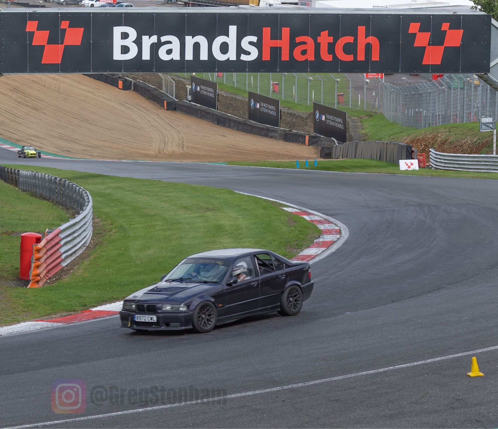 An older BMW struggling to hold its line on a wet track. The “Brands Hatch” overpass in the background, the car is exiting Druids Corner.