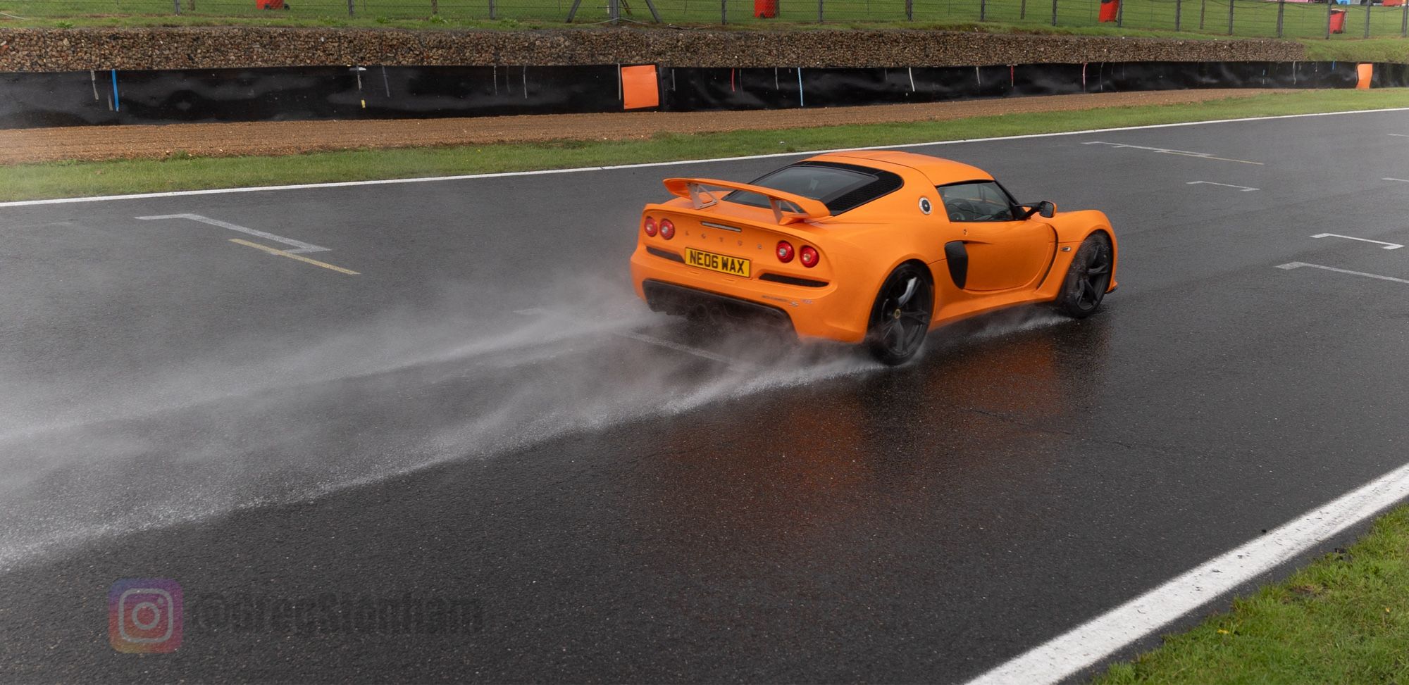 Orange coloured Lotus Exige S race car going fast down Brabham Straight at Brands Hatch. Its speed is emphasised by the water trail it’s throwing in its wake along the wet road.