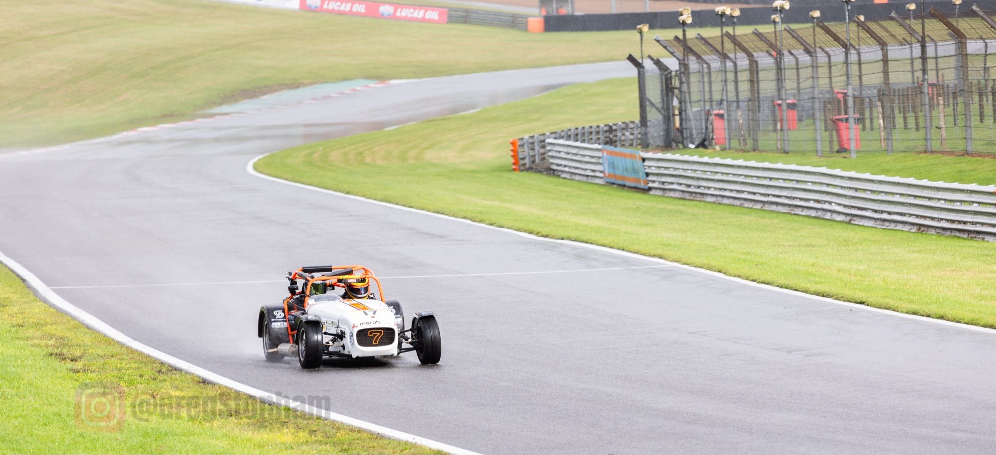 A lone, open top Caterham open top race car powering down Cooper Straight, with Graham Hill Bend in the background. Brands Hatch Circuit.