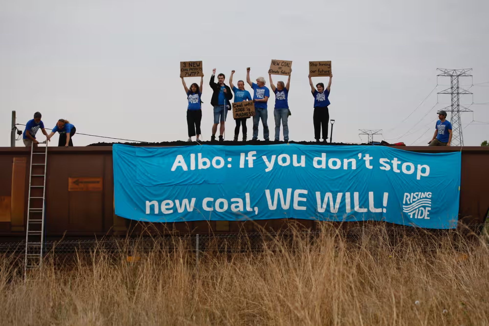 A group of people standing on top of a coal train holding up signs with one very large banner that says elbow, if you don't stop new coal, we will and the rising tide logo.