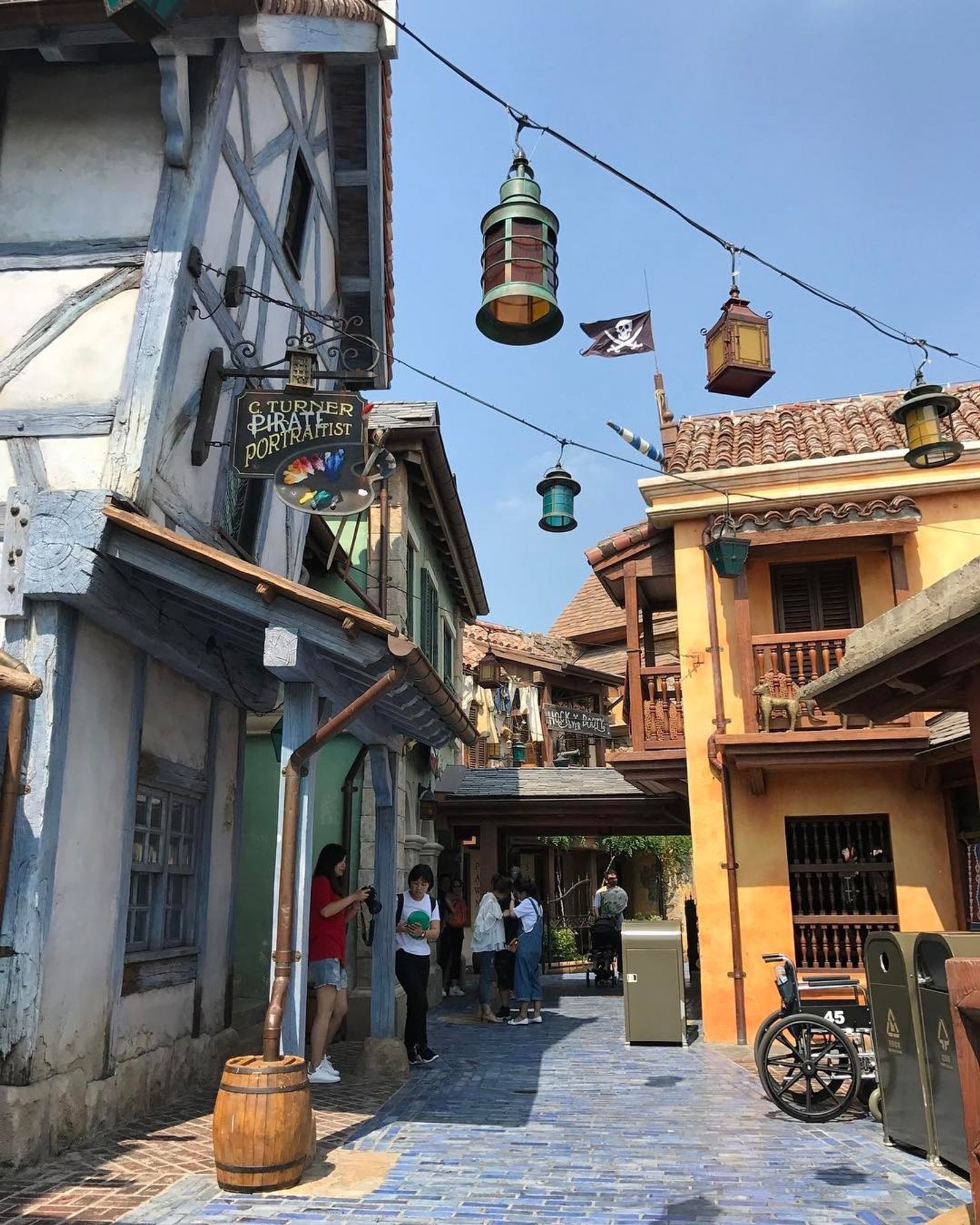 Themed alleyway with rustic buildings, hanging lanterns, trash cans, a Jolly Roger flag, and people strolling.