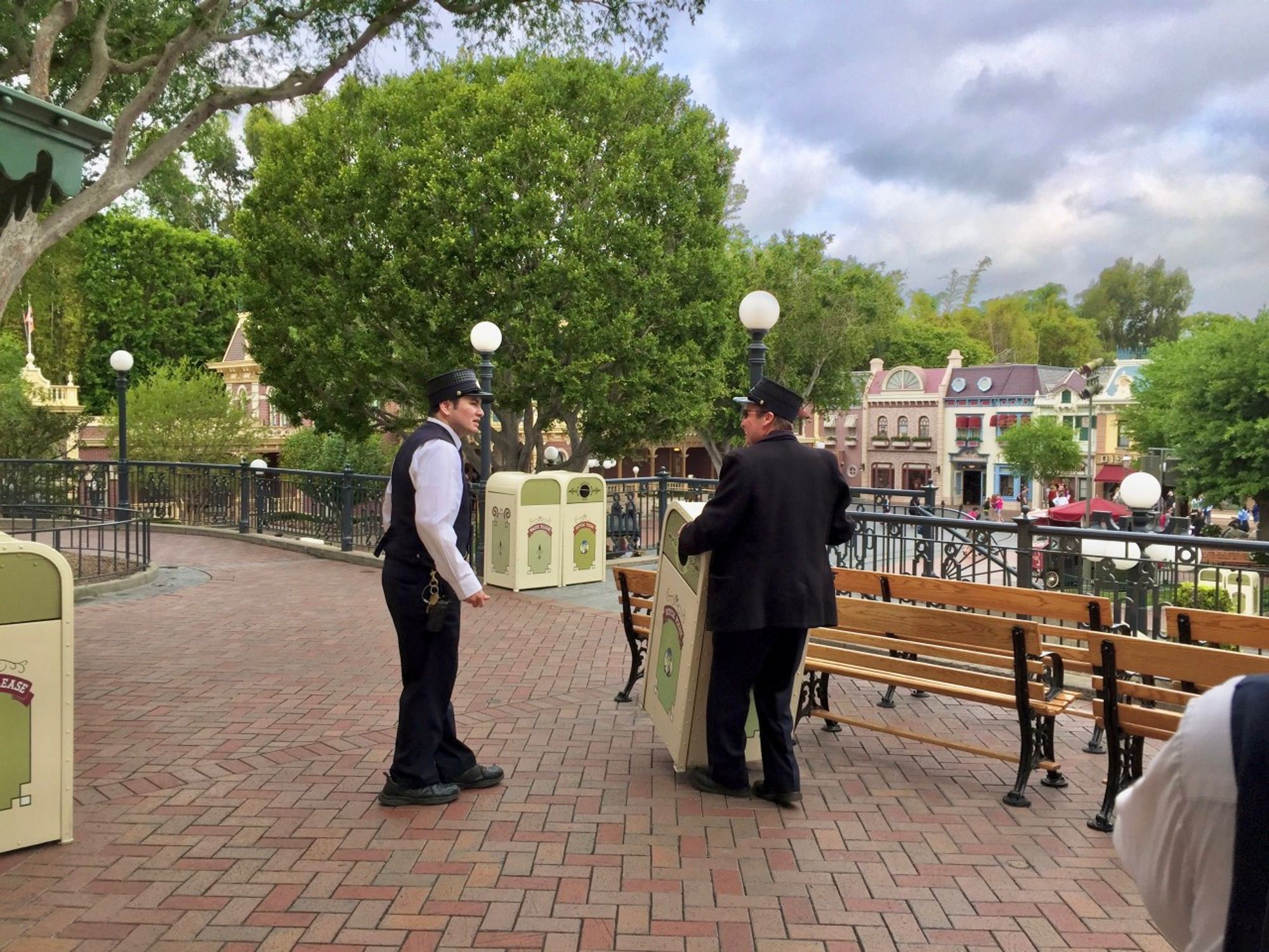 Two people in conductor uniforms standing near a bench and trash cans at a theme park, with greenery and park buildings in the background.