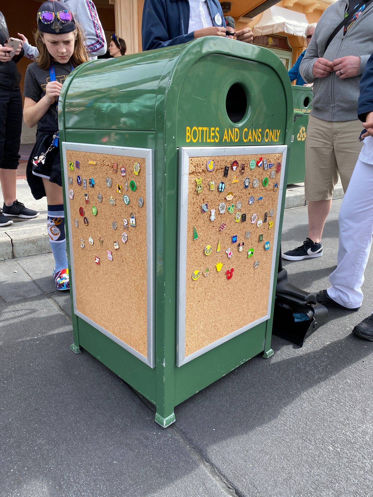 A green trash can with a corkboard displaying various pins is surrounded by several people engaged in conversation and pin trading in a park setting. Palm trees and a cloudy sky are visible in the background.