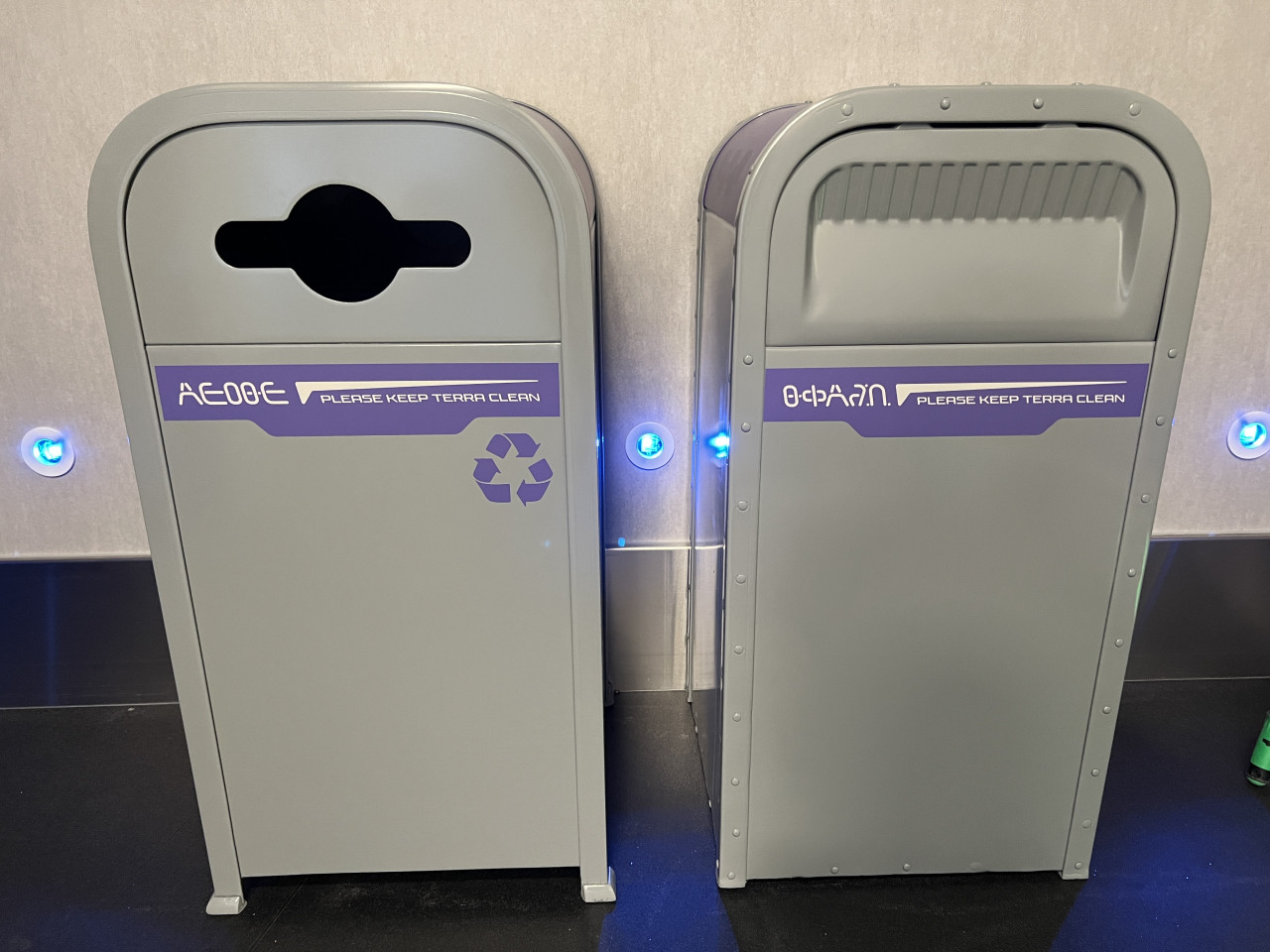 Two modern trash cans, one for recyclables with a symbol and one for trash with a push lid, both labeled "Please Keep Terra Clean" in a metallic interior with blue lights. A mysterious alien language is also featured on each can.