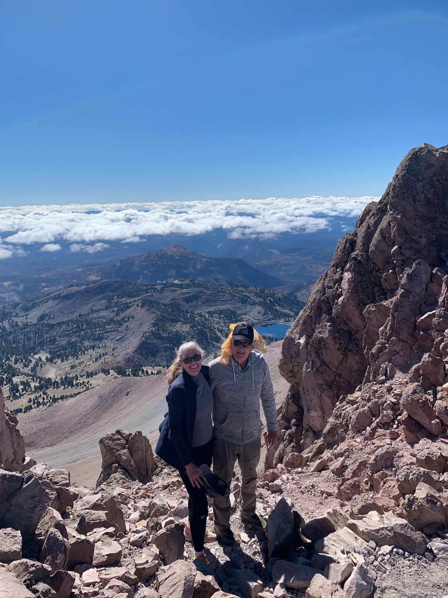 a man & a woman stand on the rock-strewn rubble of Lassen Peak.  The world below includes some clouds far off on the horizon, undulating mountains and hills, a blue lake, and the smooth sides of the volcano.