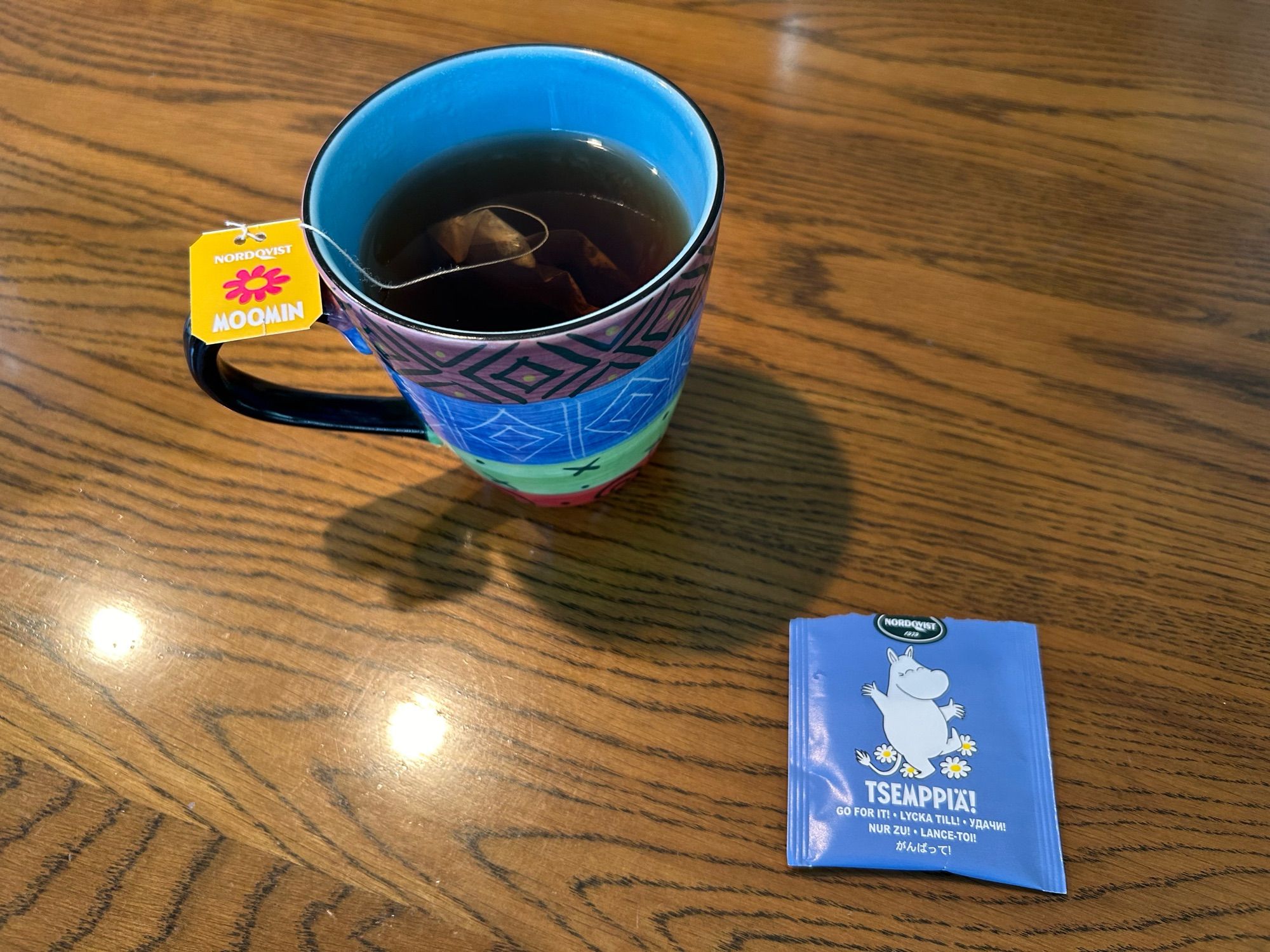 A colourful mug filled with tea sits on a wooden table, featuring a yellow tag labelled "Moomin." Next to the mug is a packet of tea with a whimsical design and the word "Tsemppiä!" printed on it.