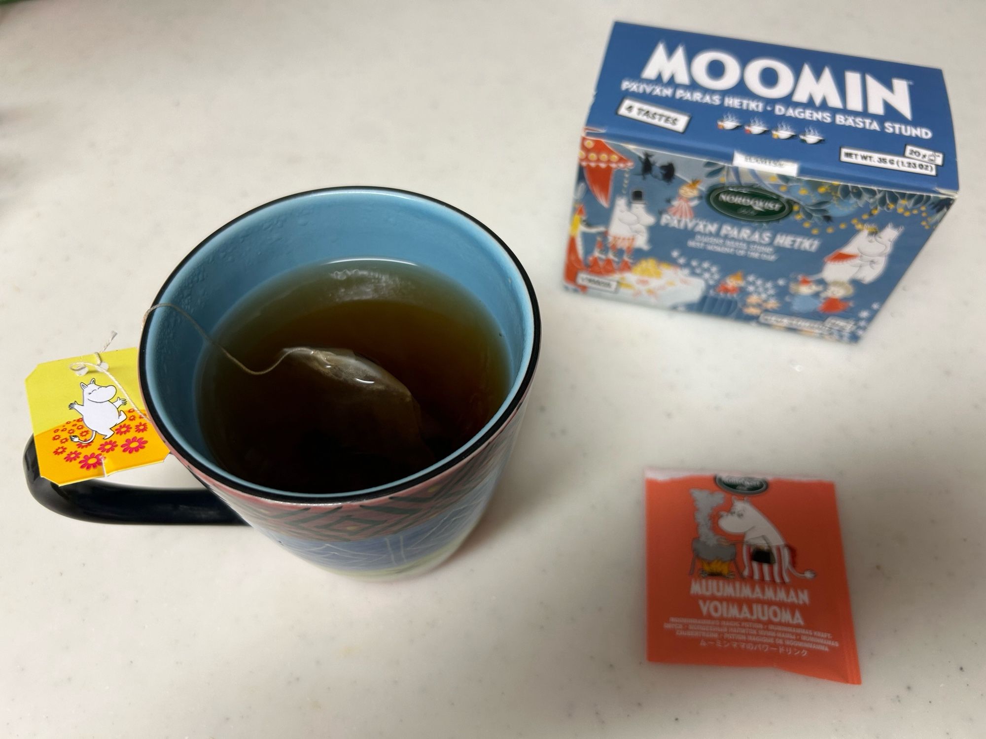 A blue mug filled with tea, teabag still steeping, is displayed on a white surface. A colourful box of Moomin-themed tea is in the background, along with an orange tea bag wrapper featuring Moominmamma.