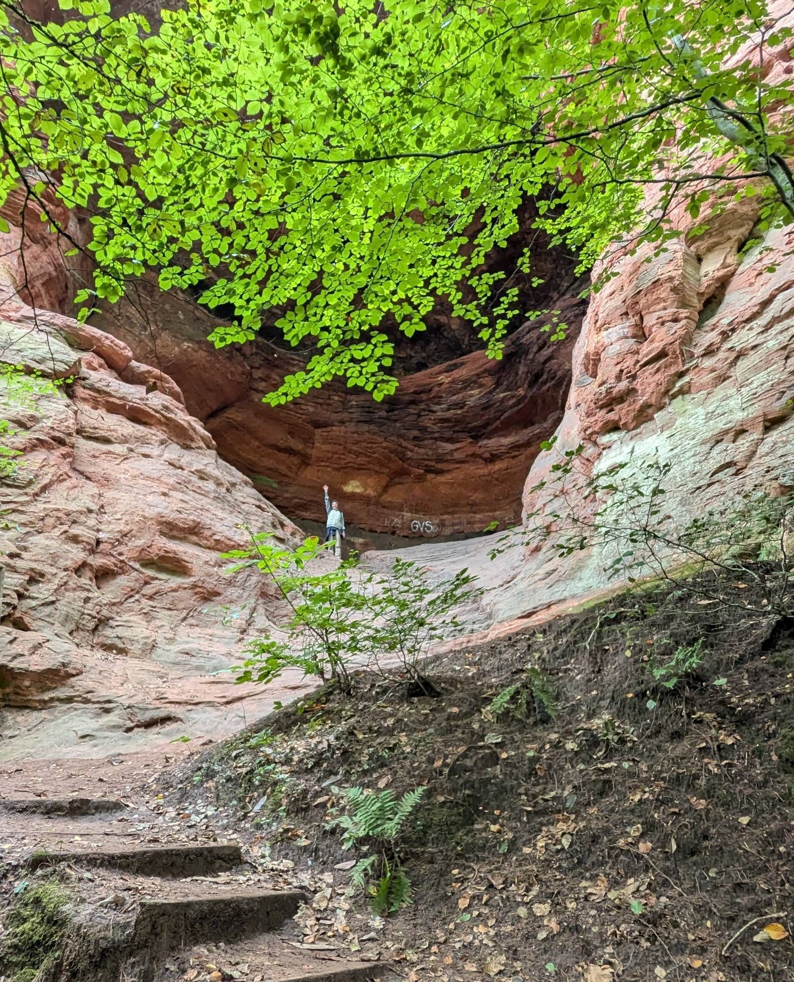 Blick von unten auf eine große Buntsandsteinhöhle, in der ganz klein ein Mensch mit hoch gerecktem Arm zu erkennen ist.