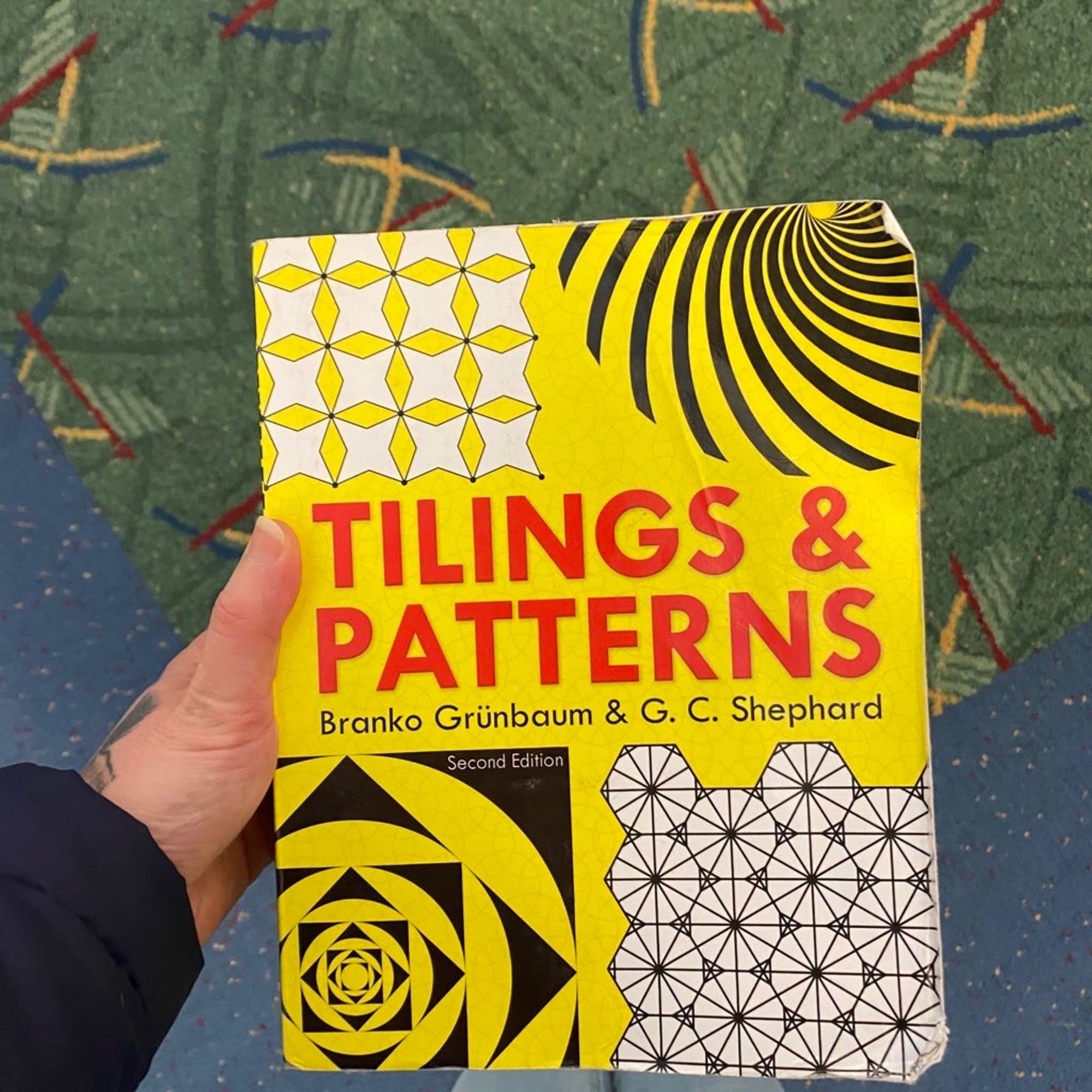 The one and only tilings and patterns by Branko Grünbaum & G.C Shephard. Dover 2ndedition, yellow background with an assortment of tilings framing the block San serif red title. This is taken at PDX and one can see a fantastic abstract deco carpet behind the book. Both this one and Gödel have been camping with me