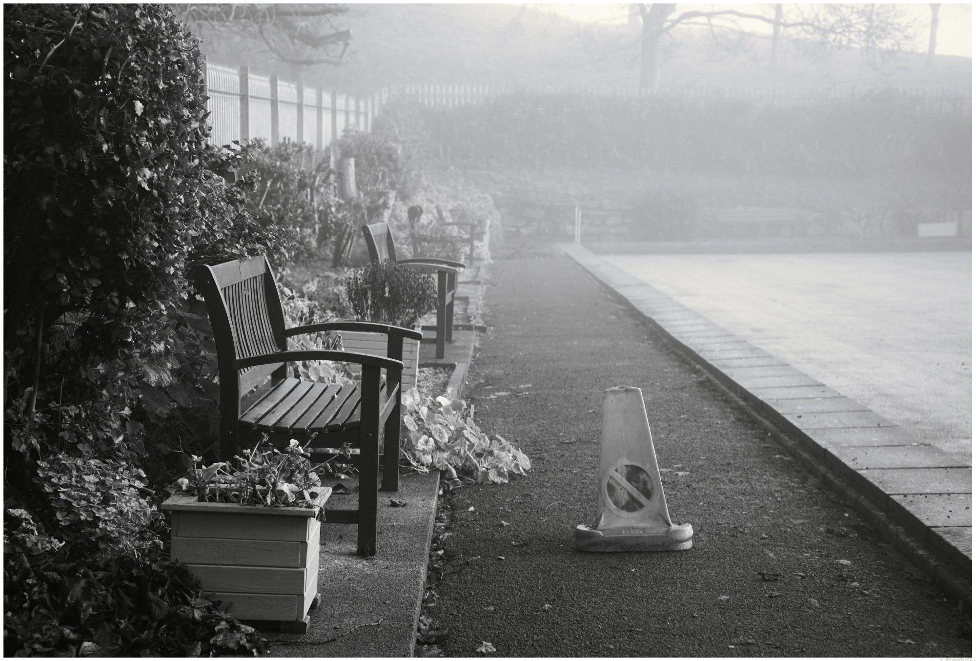 A black and white photograph of a corner of a Bowling Green, which is subject to mist hanging over the rink and away up a low hill in the backdrop.

A tarmac path runs from the lower front to the rear of the image. On the path is a repurposed 'No Entry' traffic cone.

Along the left side of the picture are four wooden, spectator seating, benches.

Nobody is playing!