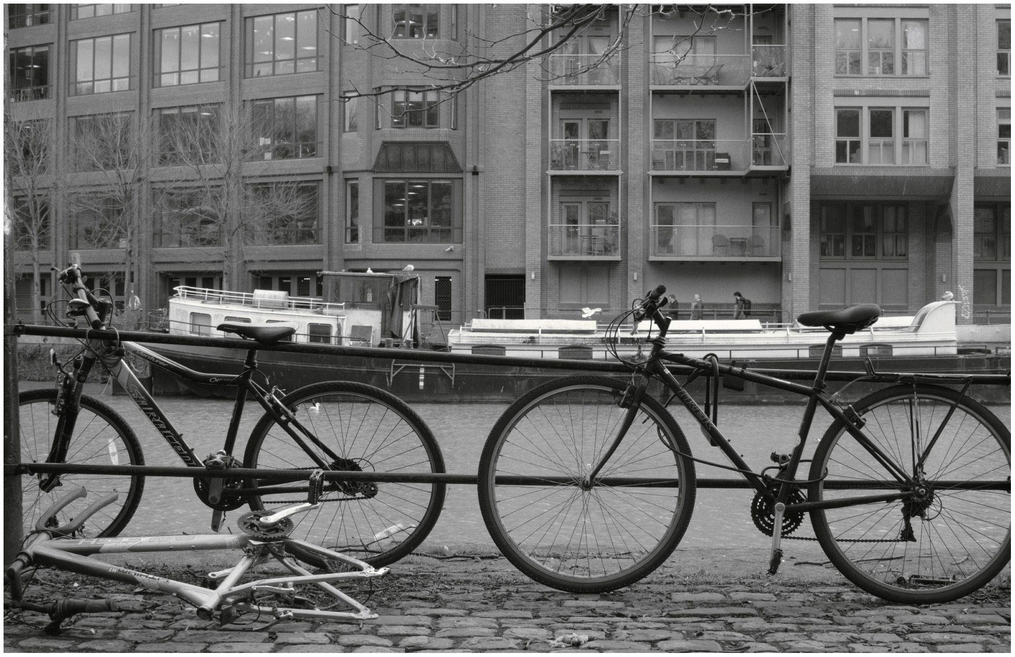 A black and white photograph taken against a backdrop of an apartment building and a barge on a river. Three bikes are shown: Two are complete and upright chained to a railing, whilst the third is without wheels and is simply a frame that has been abandoned and left on its side in the road, which is constructed of cobbles.