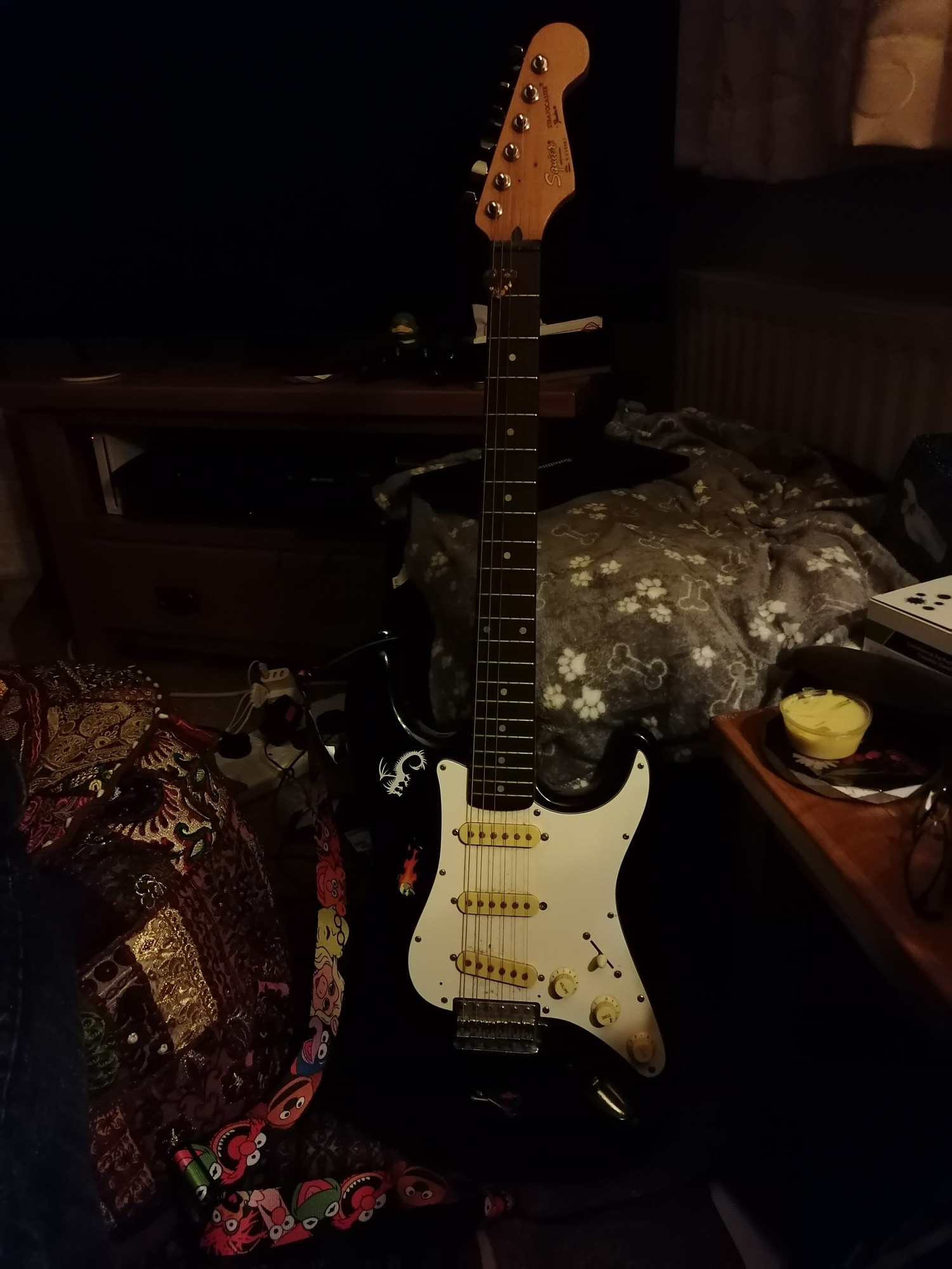 A black and white Fender Squire guitar in low light with a Muppets guitar strap.
