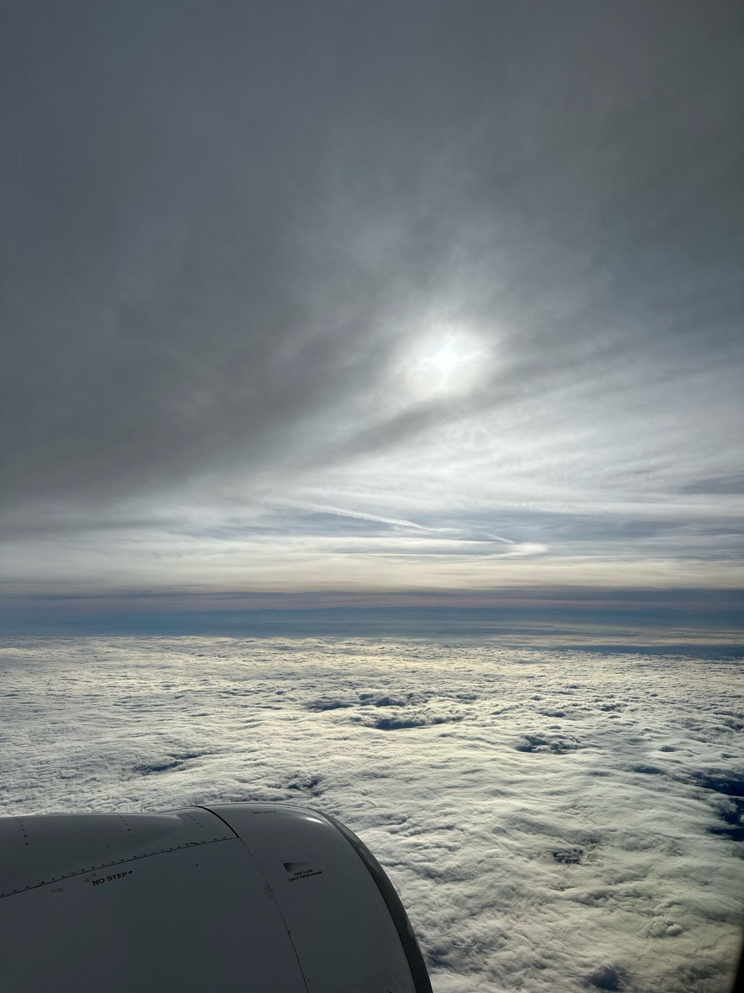 Cloud cover below with engine in the front left corner. A multitude of blue and grey colors make up the sky, with the sun barely visible behind the clouds above